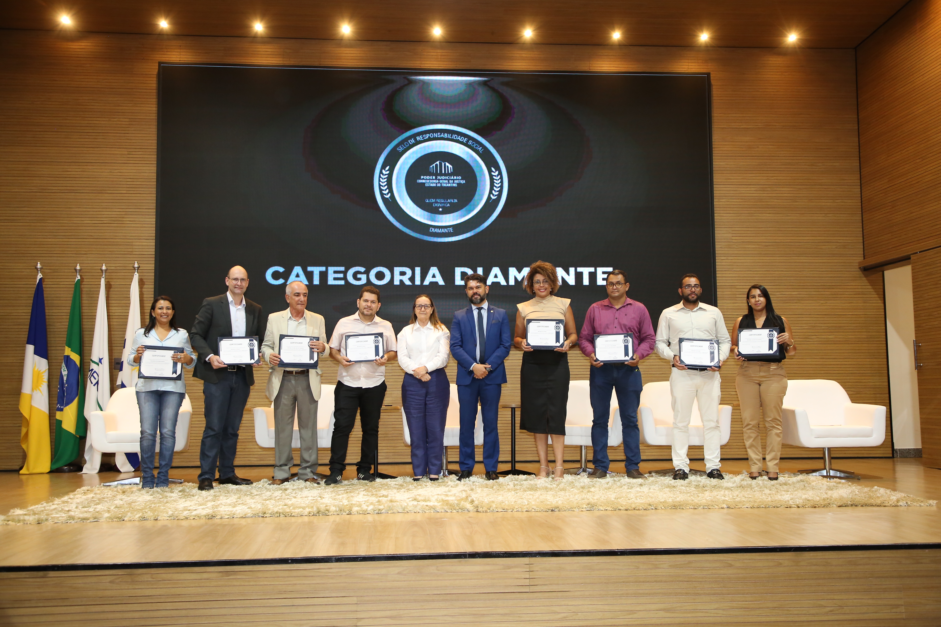 foto colorida horizontal retrada palco do auditorio do tjto onde dez pessoas, homens e mulheres posam segurando certificados