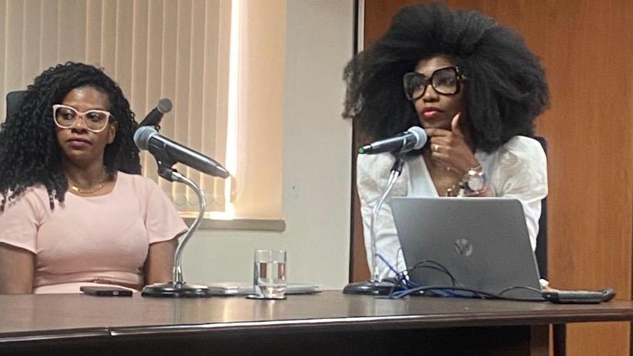 Duas mulheres negras sentadas lado a lado à mesa de madeira em tom escuro, com microfones individuais, um notebook e um copo d'água sobre a mesa. Atrás uma parede revestida de madeira e na lateral, uma parede branca com uma janela com persiana bege vertical