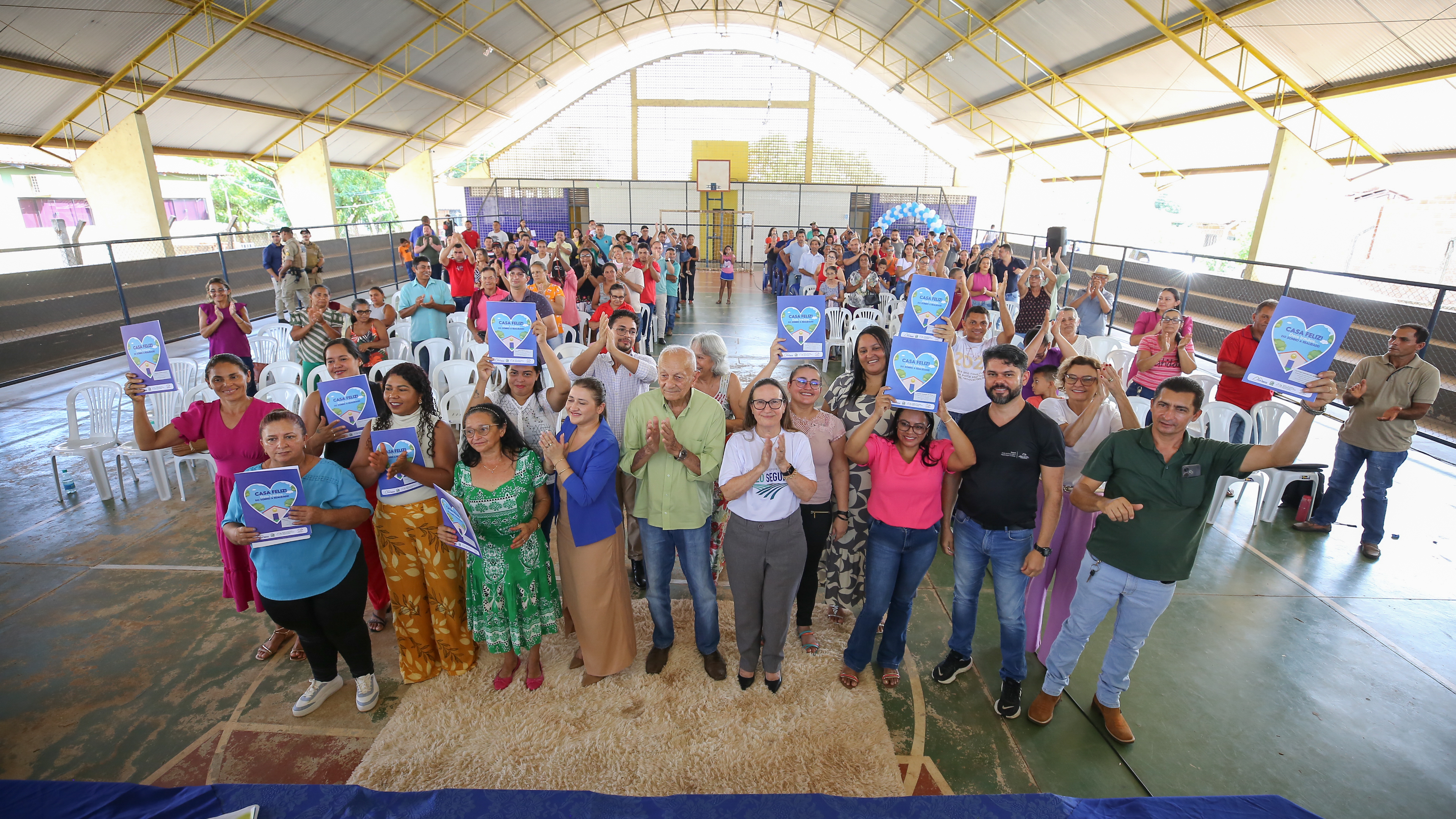 Fotografia colorida que mostra pessoas em pé, numa quadra de esportes, posando pras fotos e segurando documento.