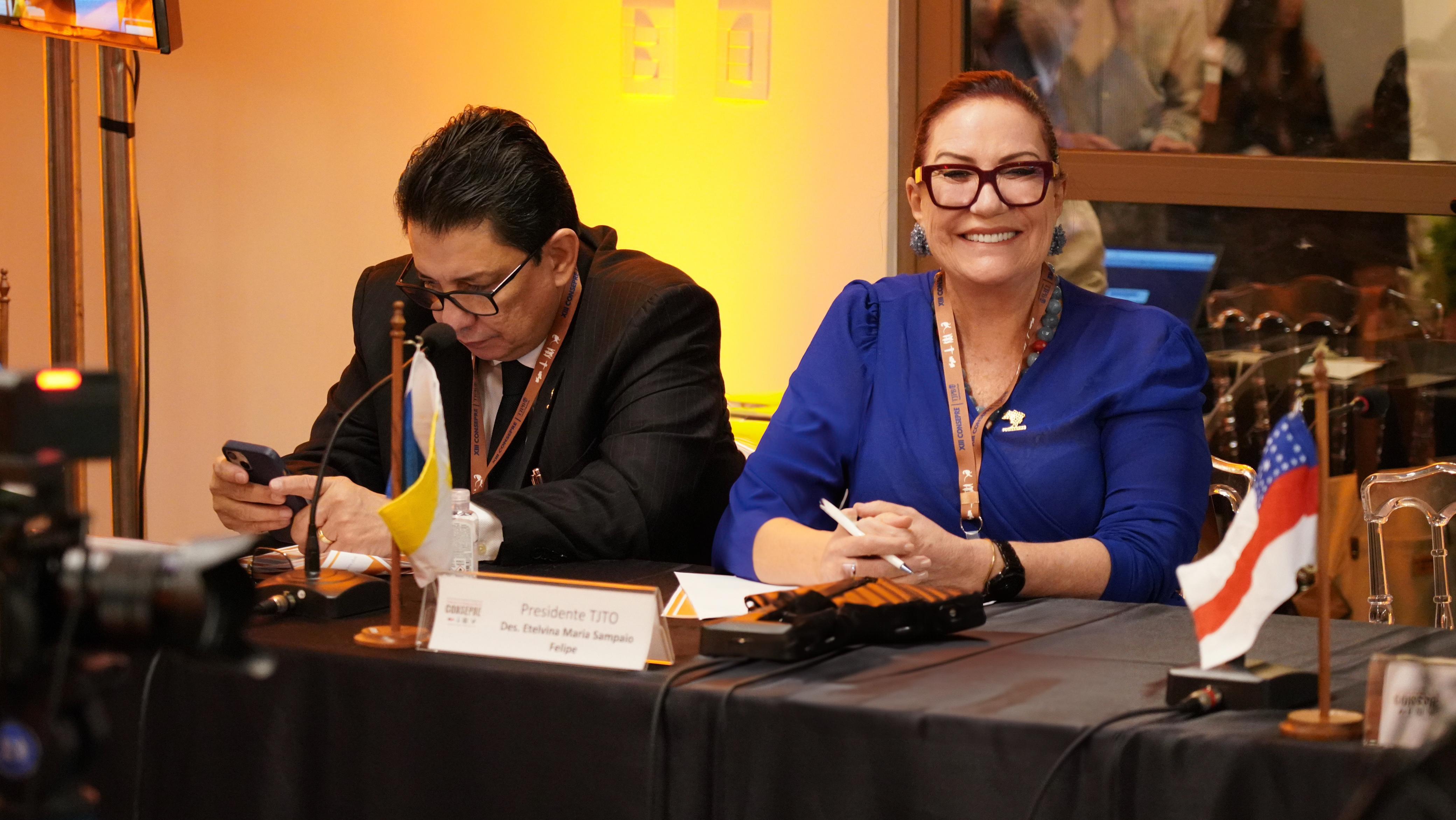 Desembargadora Etelvina, de roupa azul, aparece em primeiro plano sorrindo e posando para a foto ao lado de um outro presidente de tribunal
