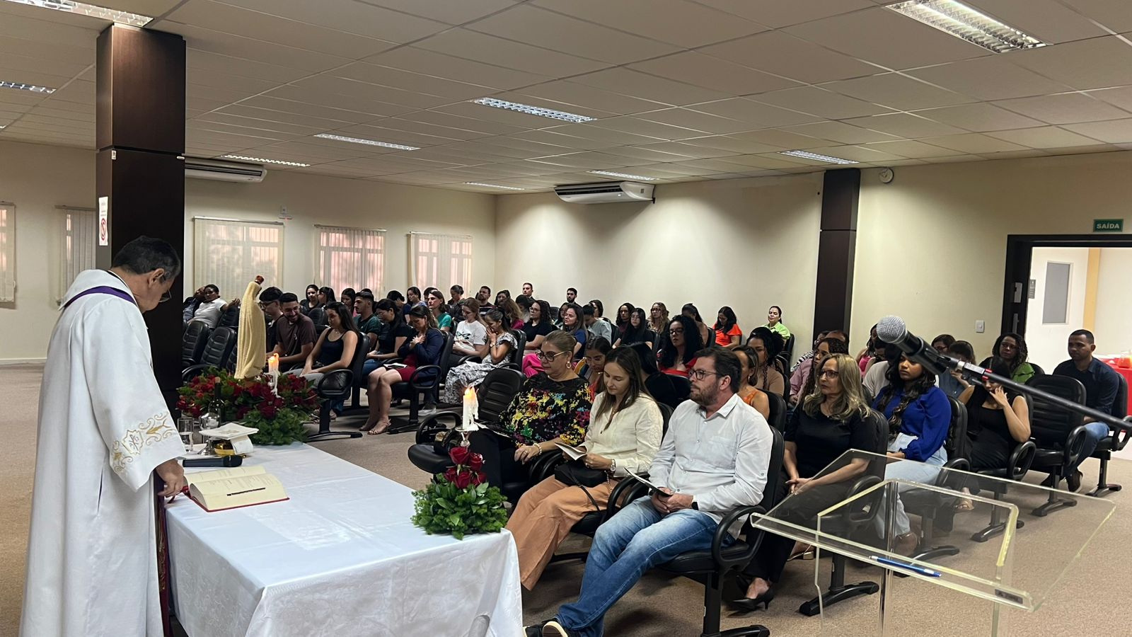fotocolorida horiontal revela a esquerda padre em pé, de cosatas, padre usando batina branca e à direita pessoas sentadas nas poltronas