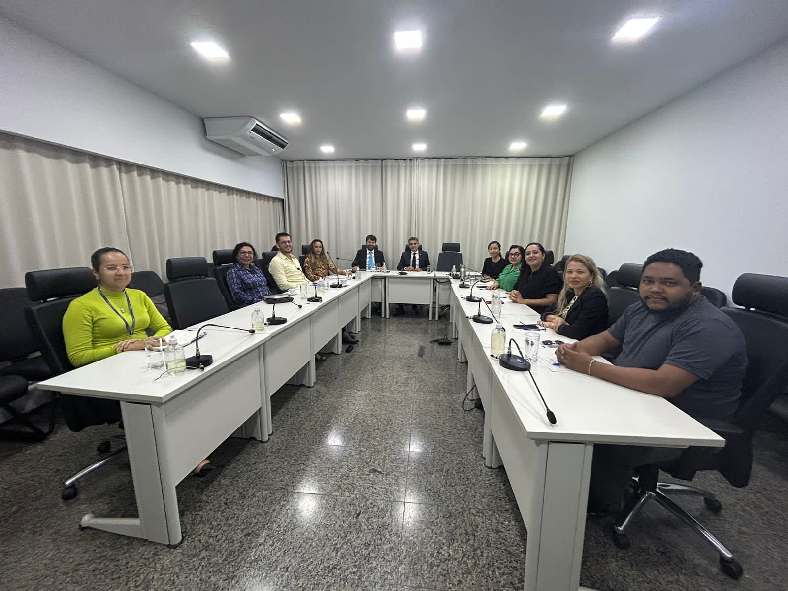 Fotografia colorida que mostra pessoas reunidas numa sala em volta de uma mesa, elas olha pra foto