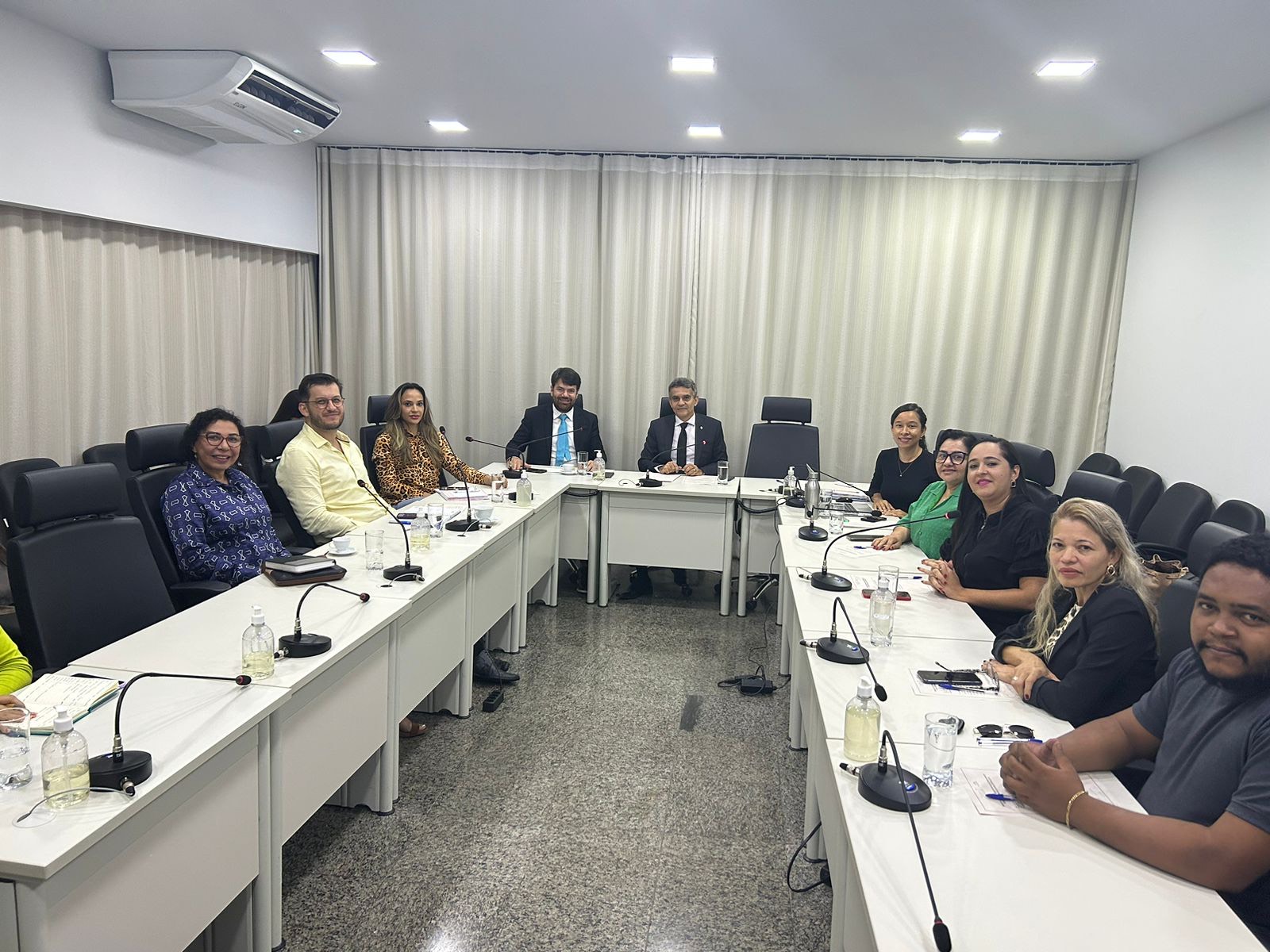 Fotografia colorida que mostra pessoas reunidas numa sala em volta de uma mesa, elas olha pra foto