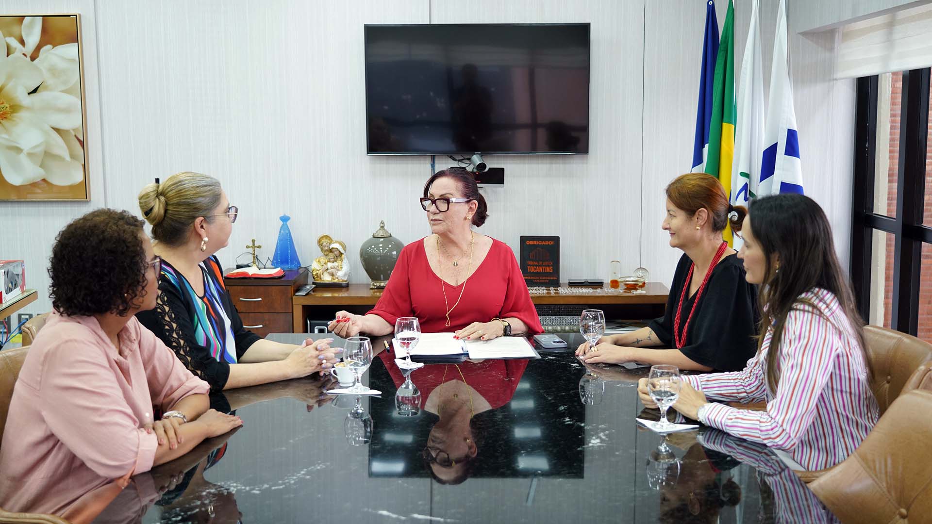 Fotografia colorida que mostra 5 mulheres sentadas à mesa. Na cabeceira está a presidente do TJTO que conversa com a juiza Flávia
