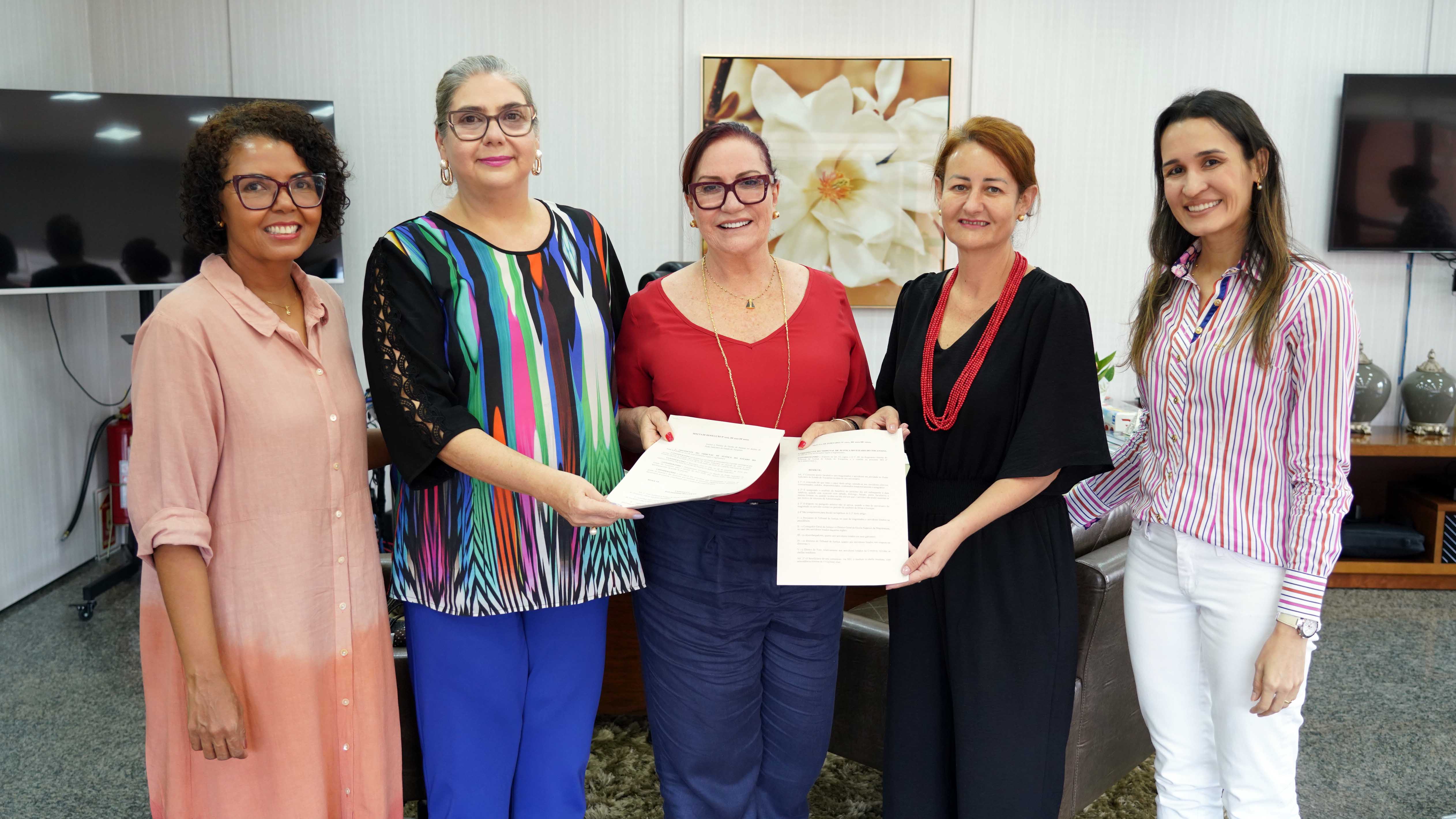 Fotografia colorida que mostra cinco mulheres sendo uma negra, posando e segurando dois documentos