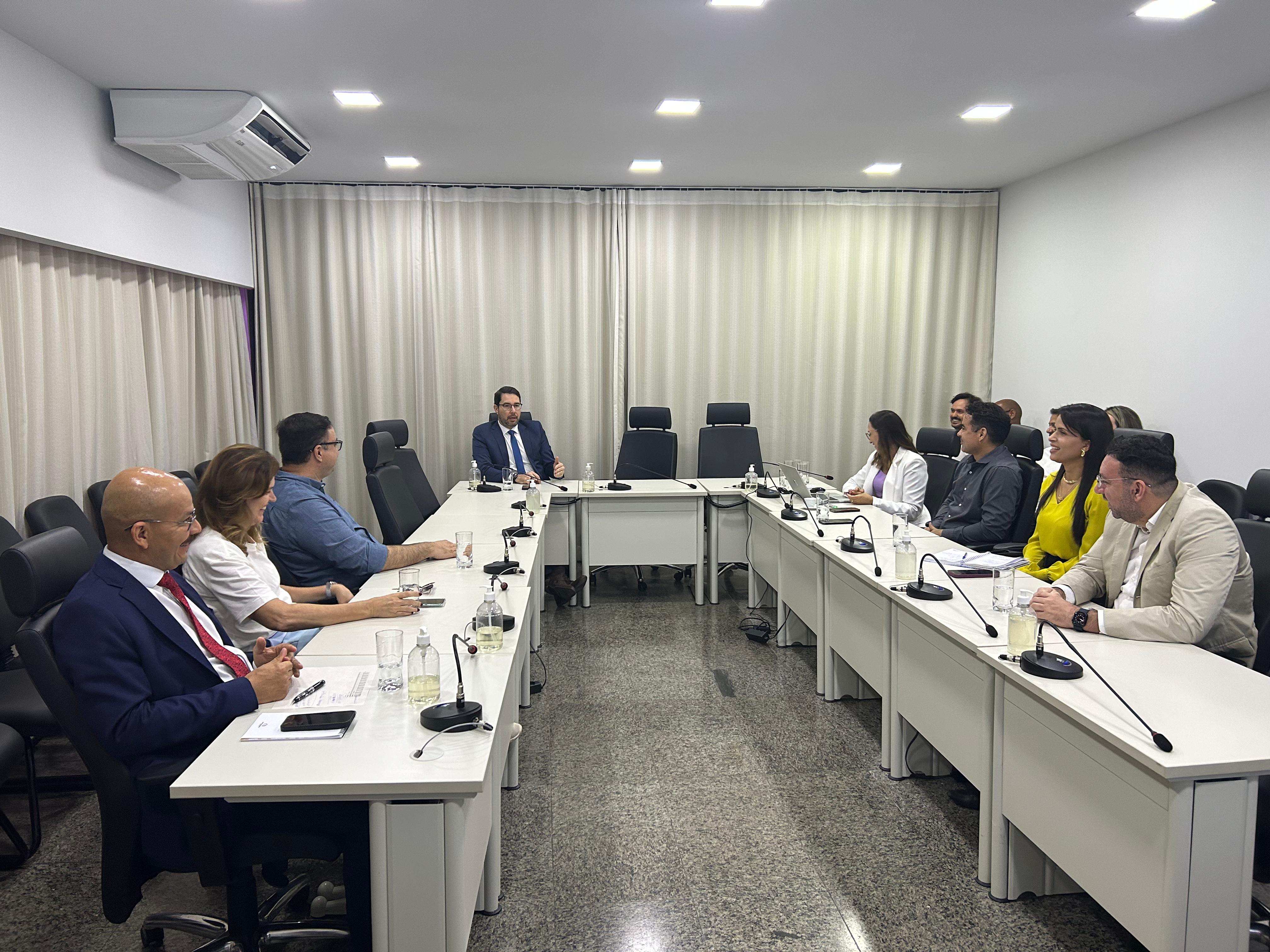 Fotografia colorida que mostra várias pessoas em uma sala de reuniões sentadas em volta da mesa