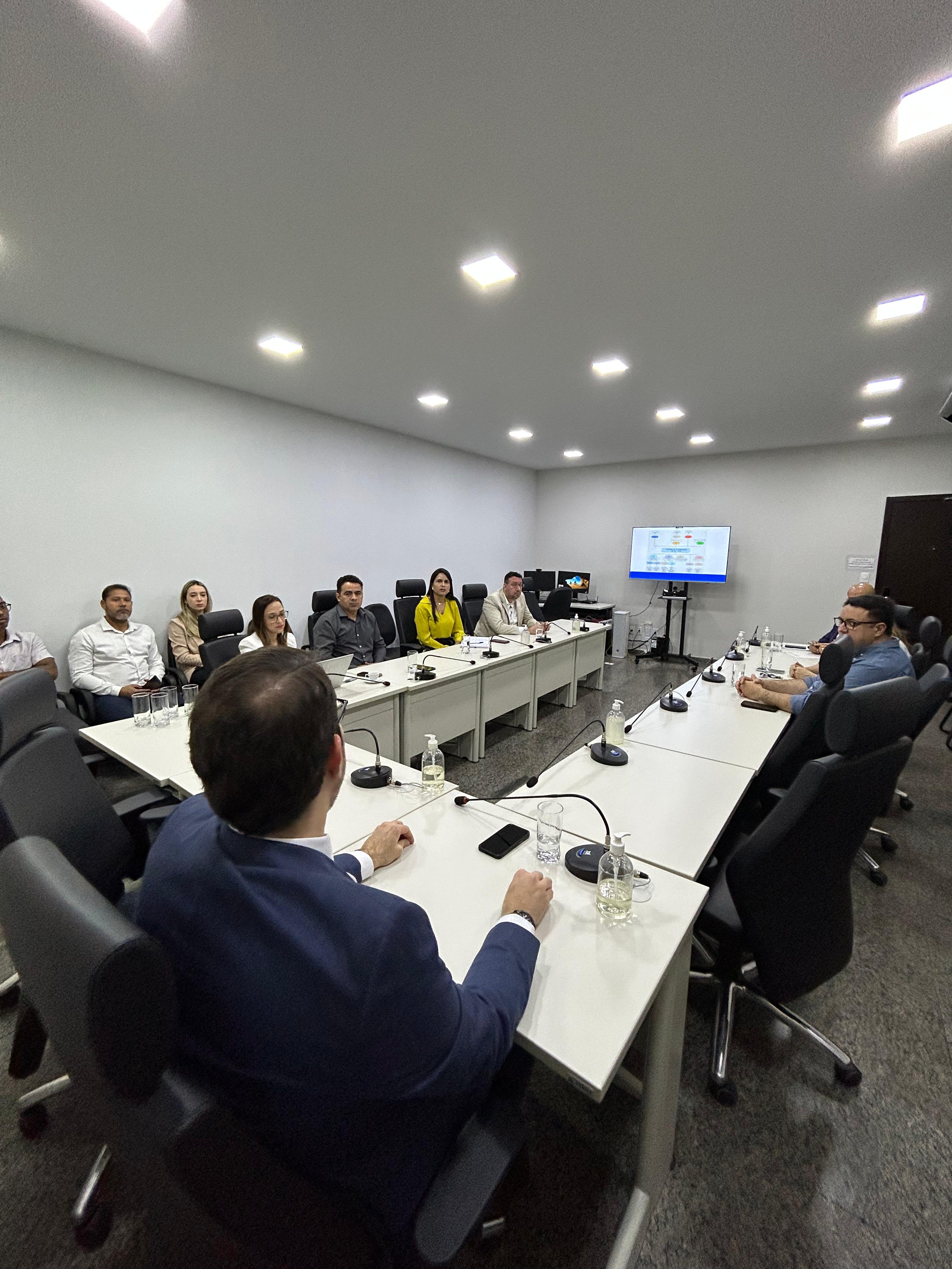 Fotografia colorida que mostra pessoas reunidas em uma sala em volta de uma grande mesa de reuniões