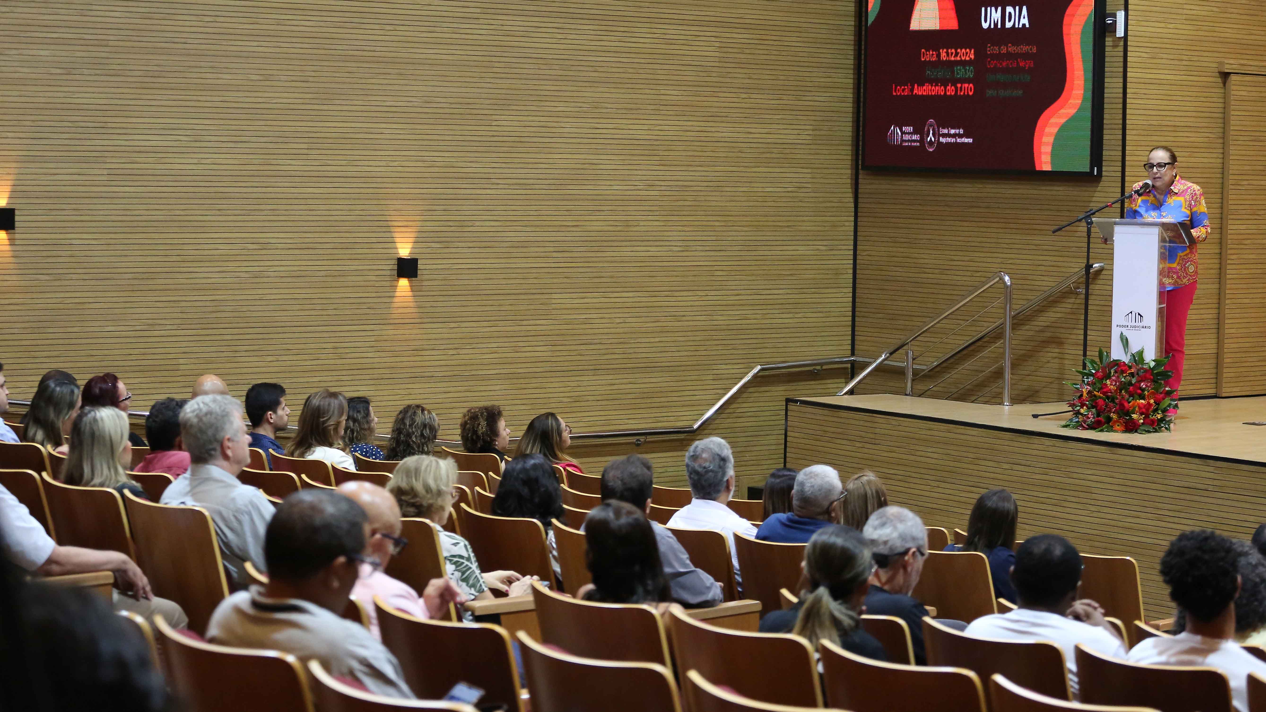 Imagem mostra uma mulher branca, em pé, com roupas coloridas falando em um púlpito dentro de um auditório em que pessoas sentadas ouvem
