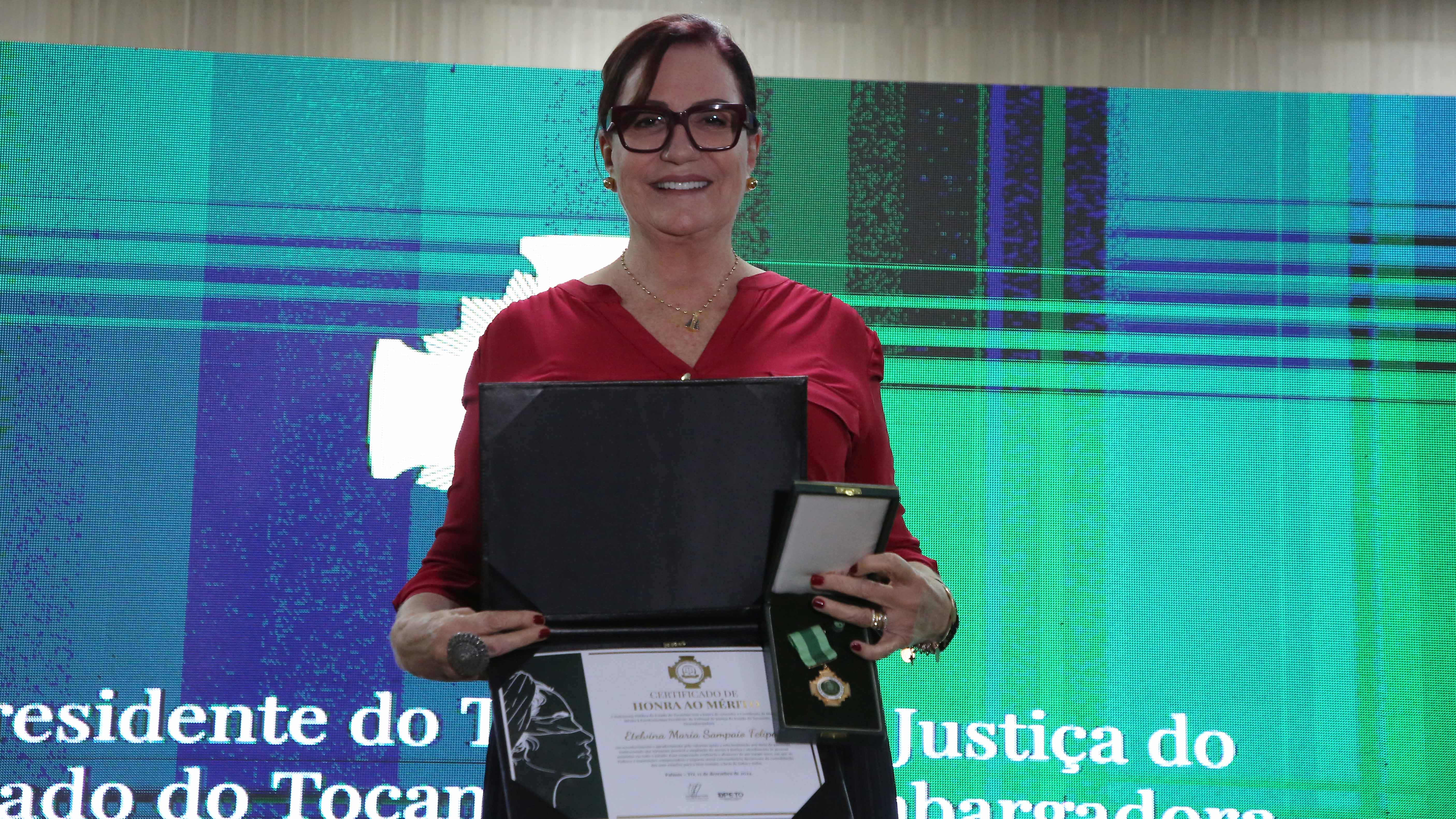 Foto da desembargadora Etelvina segurando a medalha e o diploma de Honra ao Mérito; ela está em pé, sorrindo, e usa blusa vermelha e óculos com armação escura