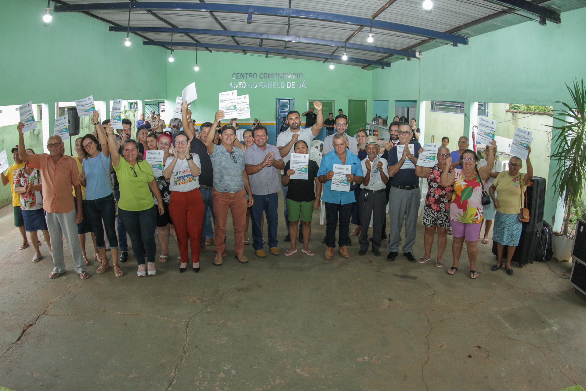 Fotografia colorida que mostra dezenas de pessoas posando para a foto e segurando um documento de regularização fundiária. Eles estão em um galpão aberto
