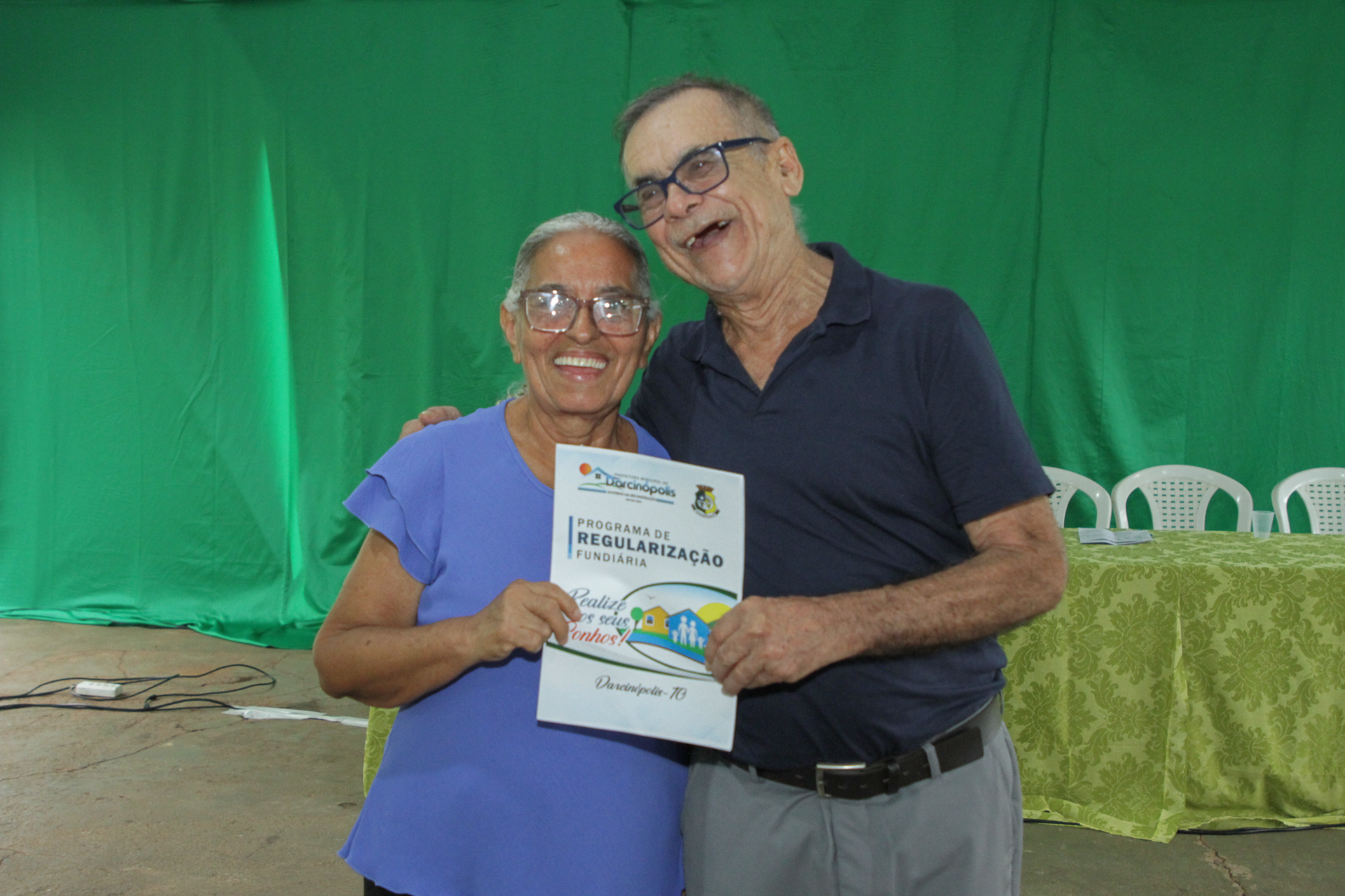 Imagem colorida que mostra uma senhora sorridente recebendo um titulo de propriedade de um homem grisalho