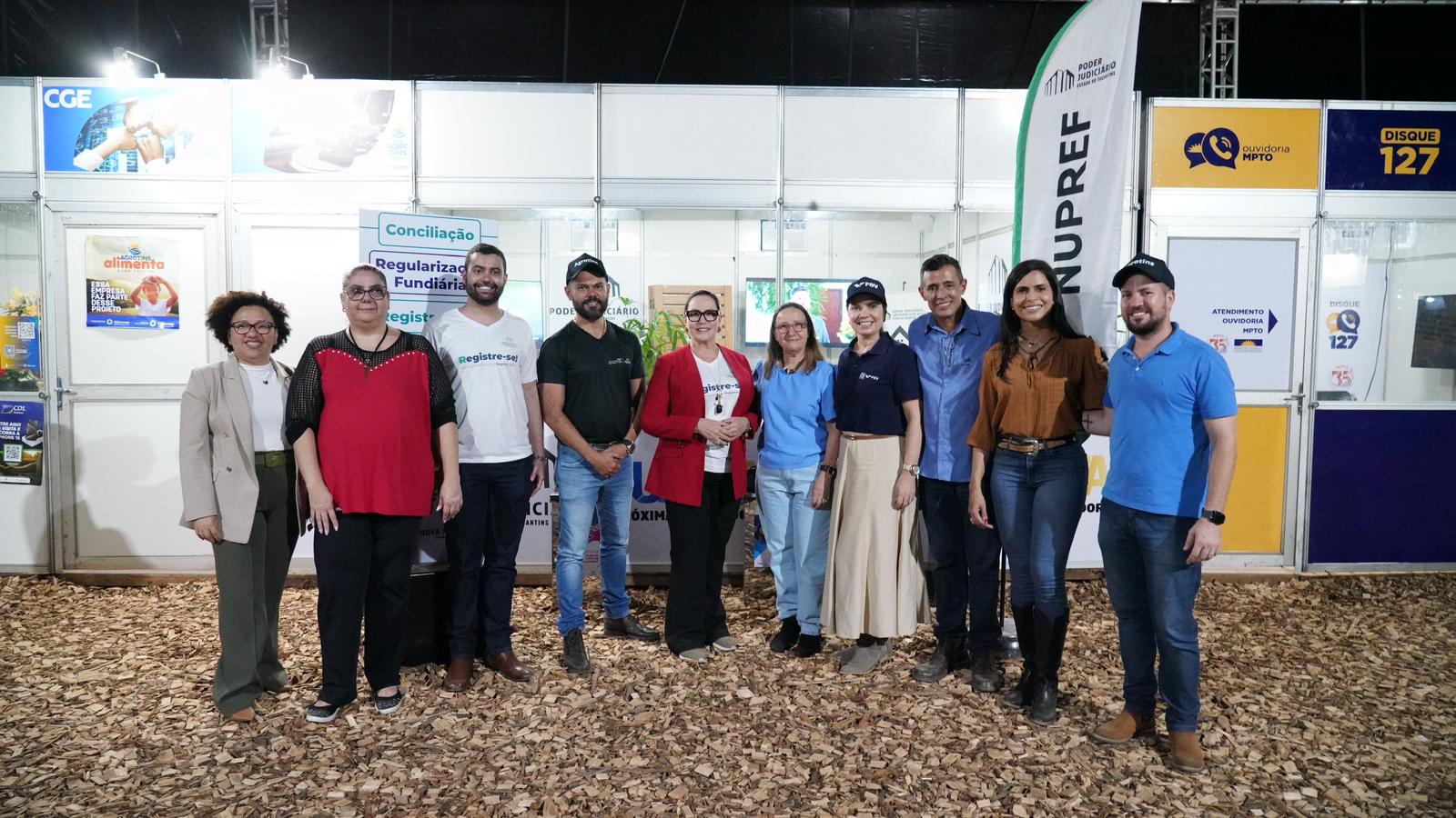 Fotografia colorida onde dez pessoas, seis mulheres e quatro homens, posam para foto lado a lado em frente a estandes da agrotins 2024