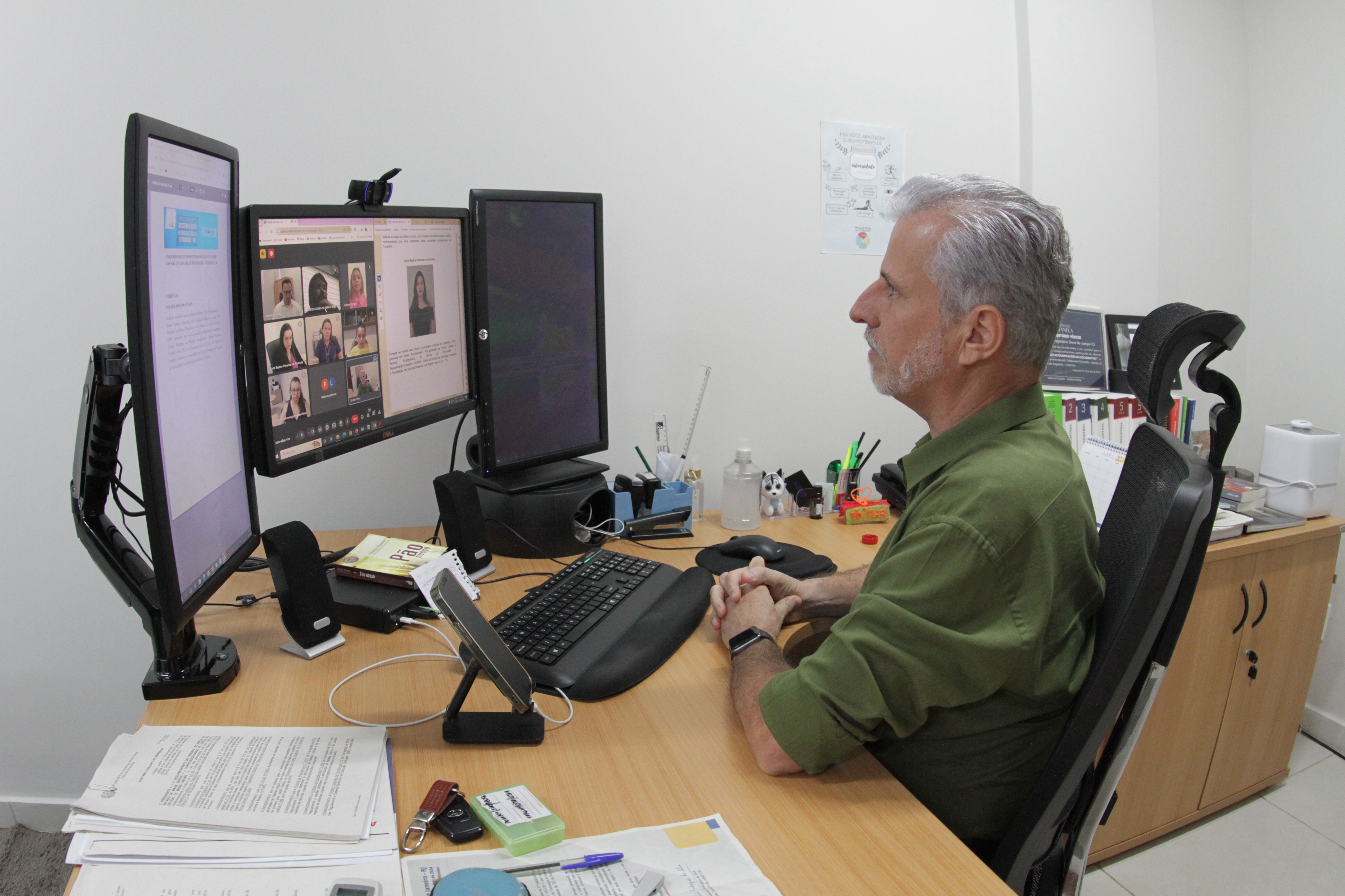 Fotografia colorida onde um senhor branco, de camisa verde e cabelos e barba grisalhos está sentado em uma cadeira em frente a um computador, que possui três telas, em cima de uma mesa de madeira clara com diversos objetos como celular, canetas e papeis 