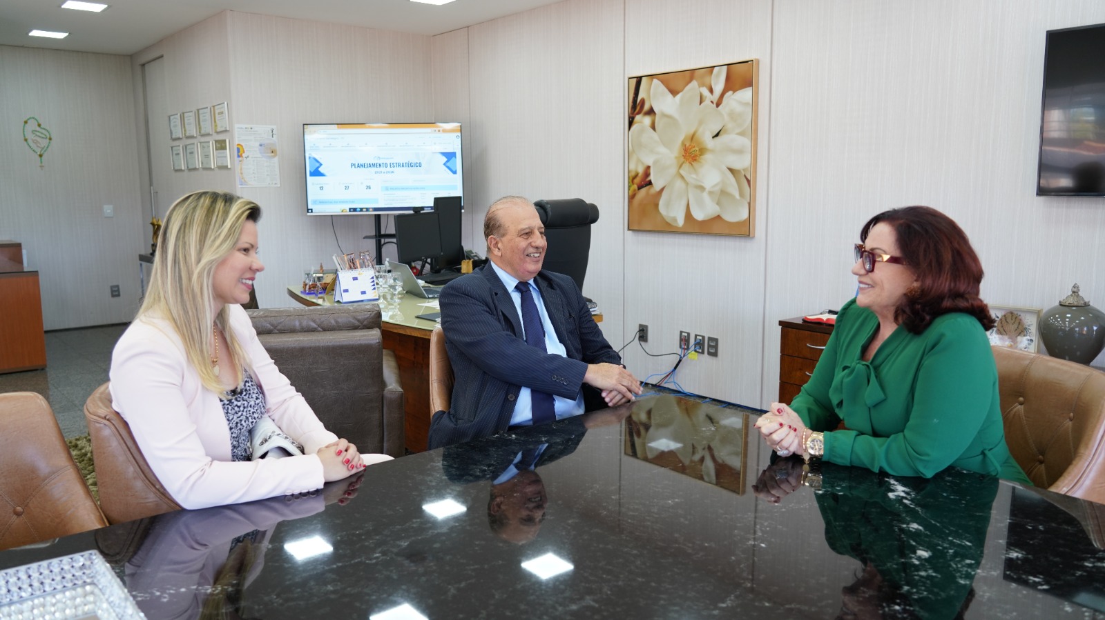 Três pessoas sentadas ao redor da mesa. Sendo elas duas mulheres, uma ruiva, uma loira um homem de terno e gravata.