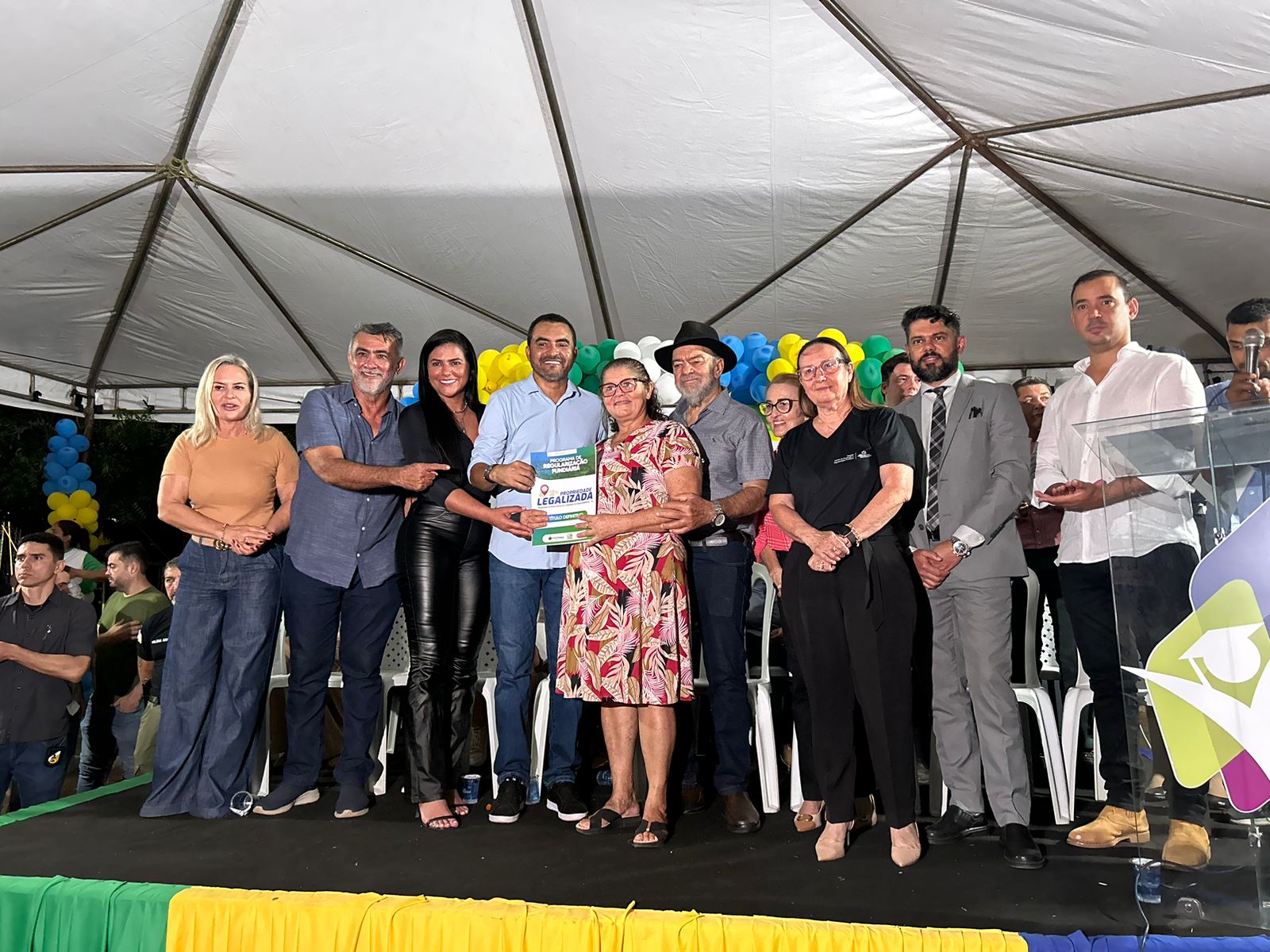 Fotografia colorida de onze pessoas posando para fotografia uns dos lados dos outros, em cima de um palco preto decorado com balões e panos verde e amarelos em volta. Eles estão em baixo de uma tenda de lona branca e uma senhora de vestido com detalhes de flores vermelhos, brancos e amarelos, que usa óculos, segura um documento de regularização fundiária