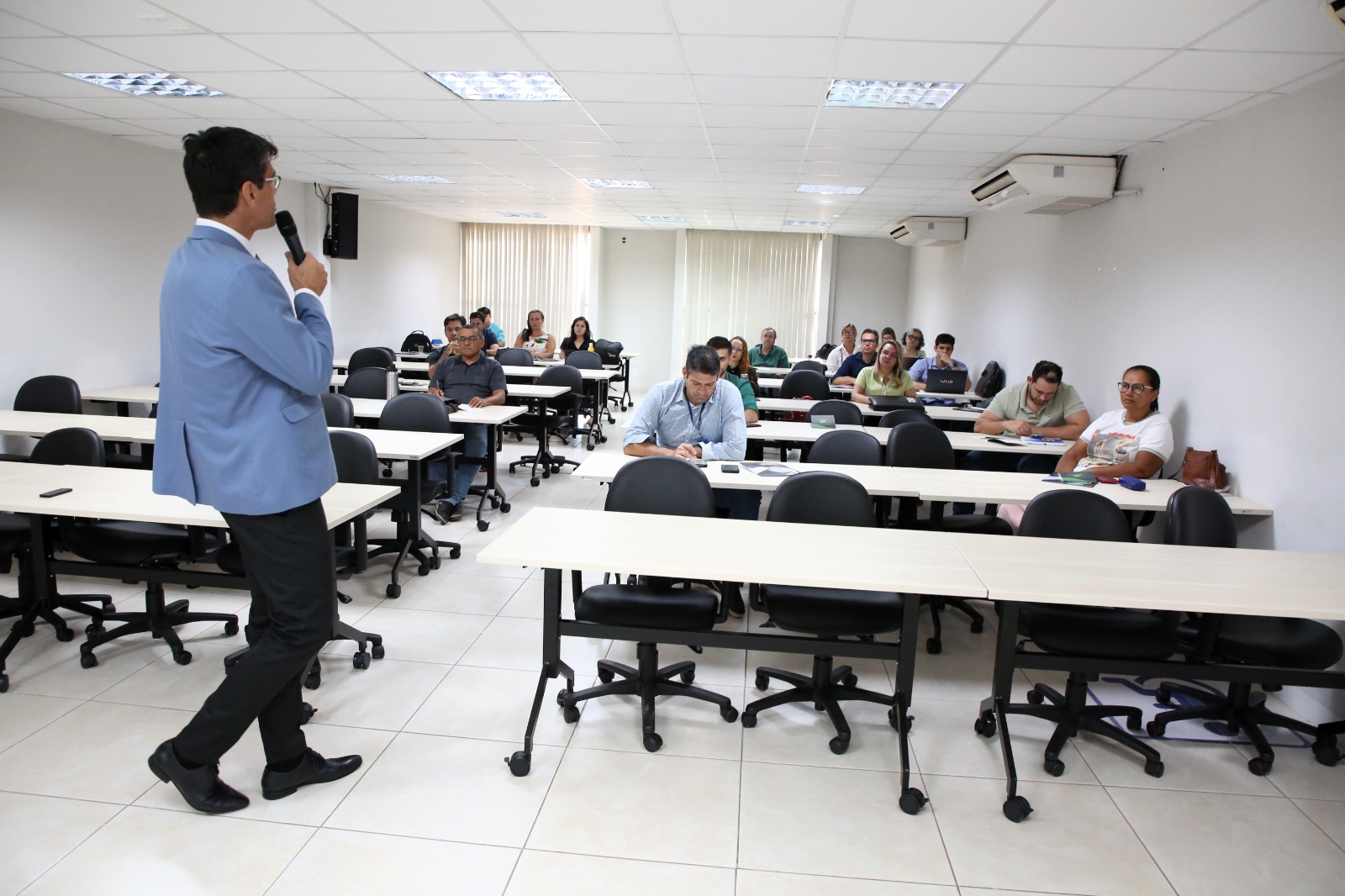 Fotografia colorida onde um homem está de pé falando em um microfone para um grupo de pessoas que estão sentados em sua frente. Eles estão em uma sala branca.