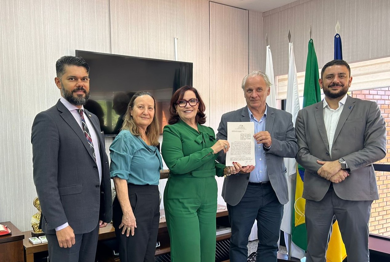 Fotografia colorida onde a presidente do Tribunal de Justiça do Tocantins até no centro, de roupa verde, óculos e cabelo solto, segurando um documento junto de um homem com terno cinza, camisa branca por dentro, branco de cabelos grisalhos. Ao lado deles estão mais uma mulher e dois homens. Ao fundo, se encontra um painel de madeira clara, com uma televisão e quatro bandeiras.