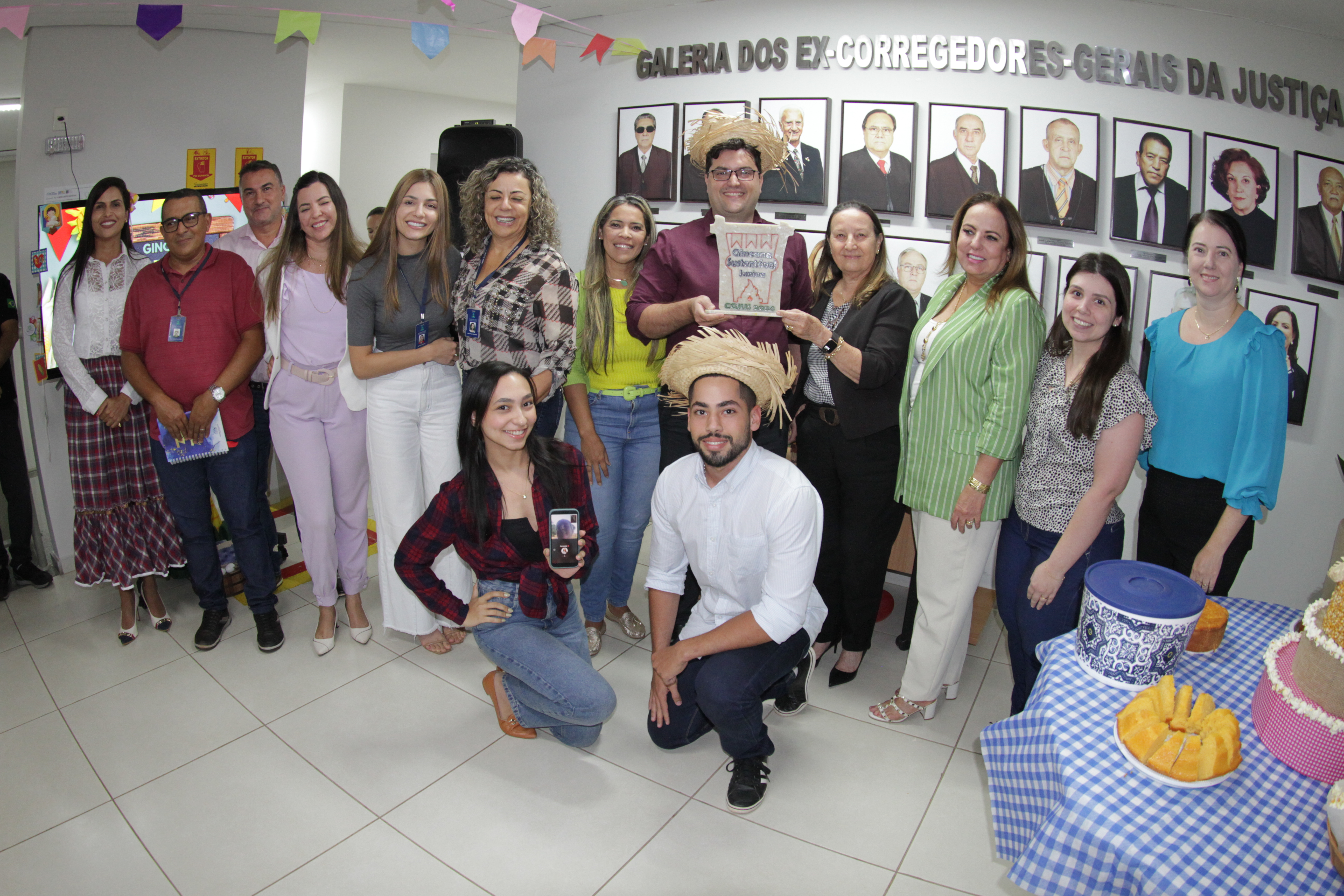 Fotografia colorida onde um grupo de pessoas está posando para foto caracterizadas com roupas temáticas de festa junina em frente a uma parede com fotos de vários corregedores dentro de uma sala branca 
