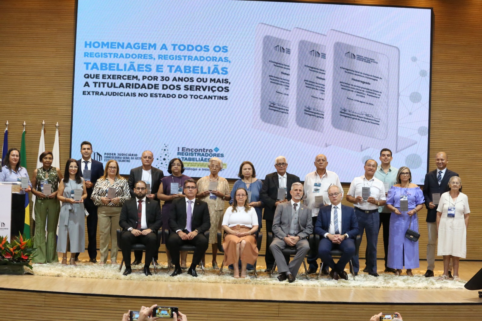 Grupo de pessoas estão posicionadas tanto em pé quanto sentadas e seguram placas indicando que estão sendo reconhecidas.