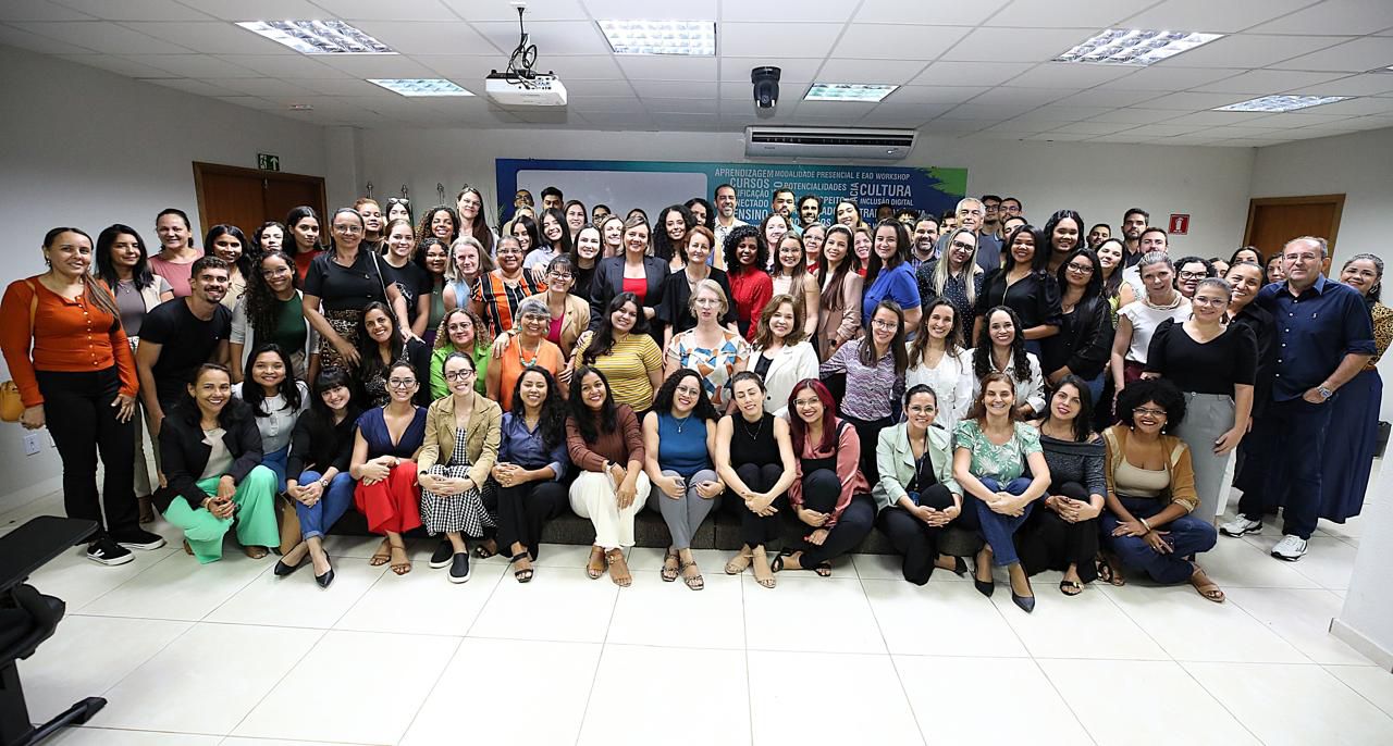foto colorida horizontal mostra mulheres e homens, alguns sentados, outros de pé pousando para foto. eles estao em uma sala