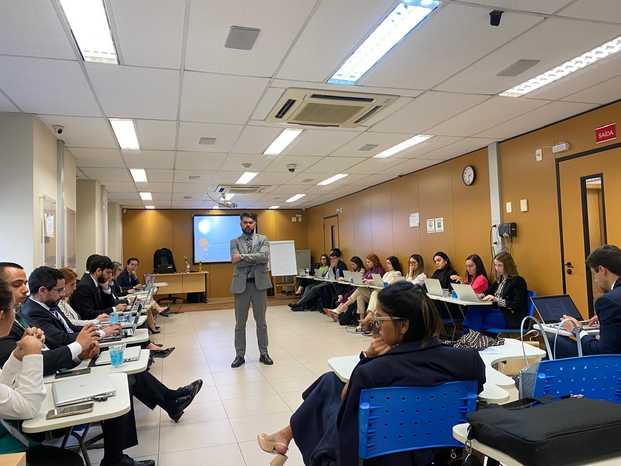 Fotografia colorida onde um homem de terno e calça cinzas está parado no meio de uma sala, de braços cruzados, com várias pessoas sentadas em cadeiras a sua volta, e ao fundo uma mesa de madeira clara e uma grande tela 