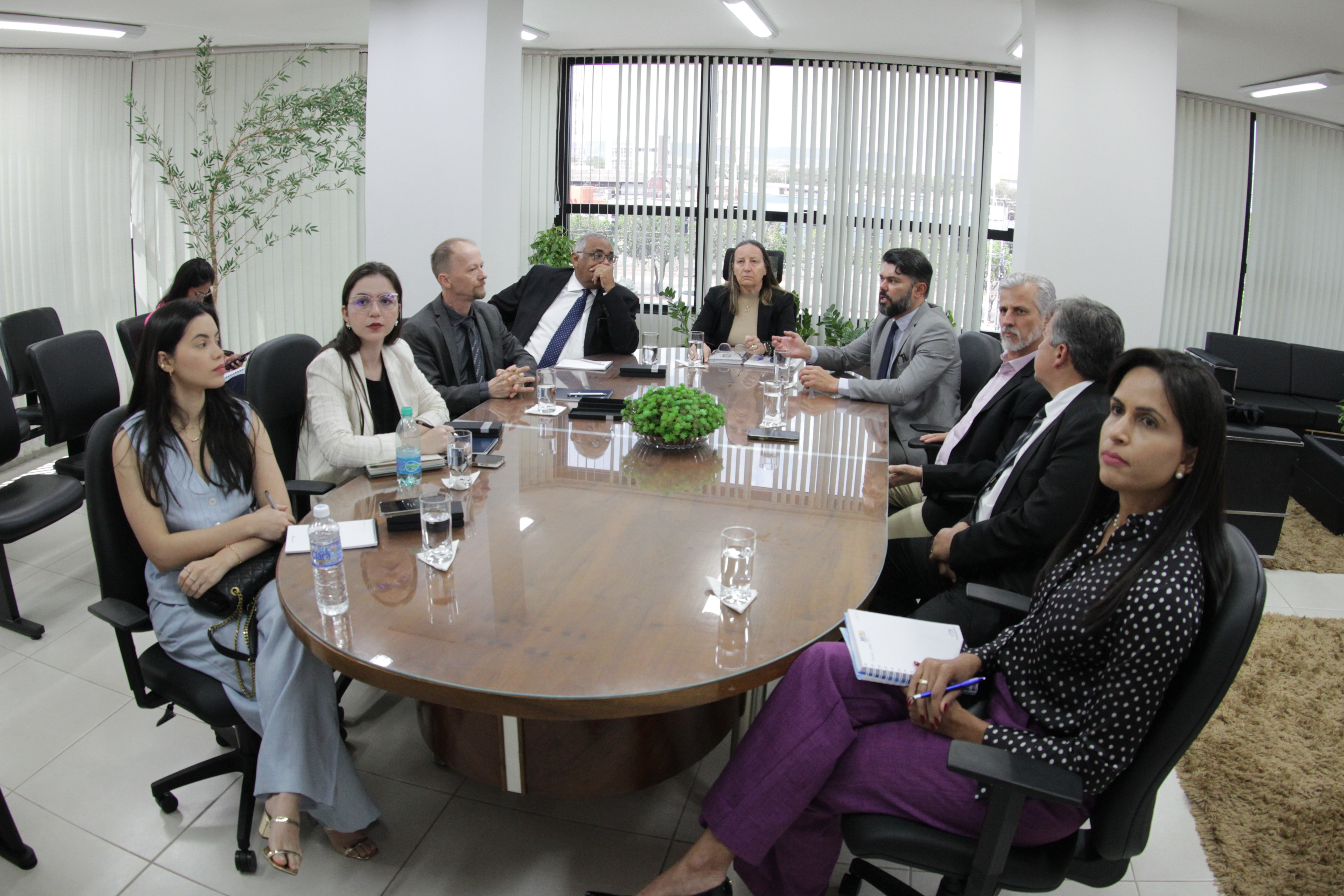 Nove pessoas sentadas ao redor de uma mesa oval de reunião, sendo quatro mulheres e cinco homens.