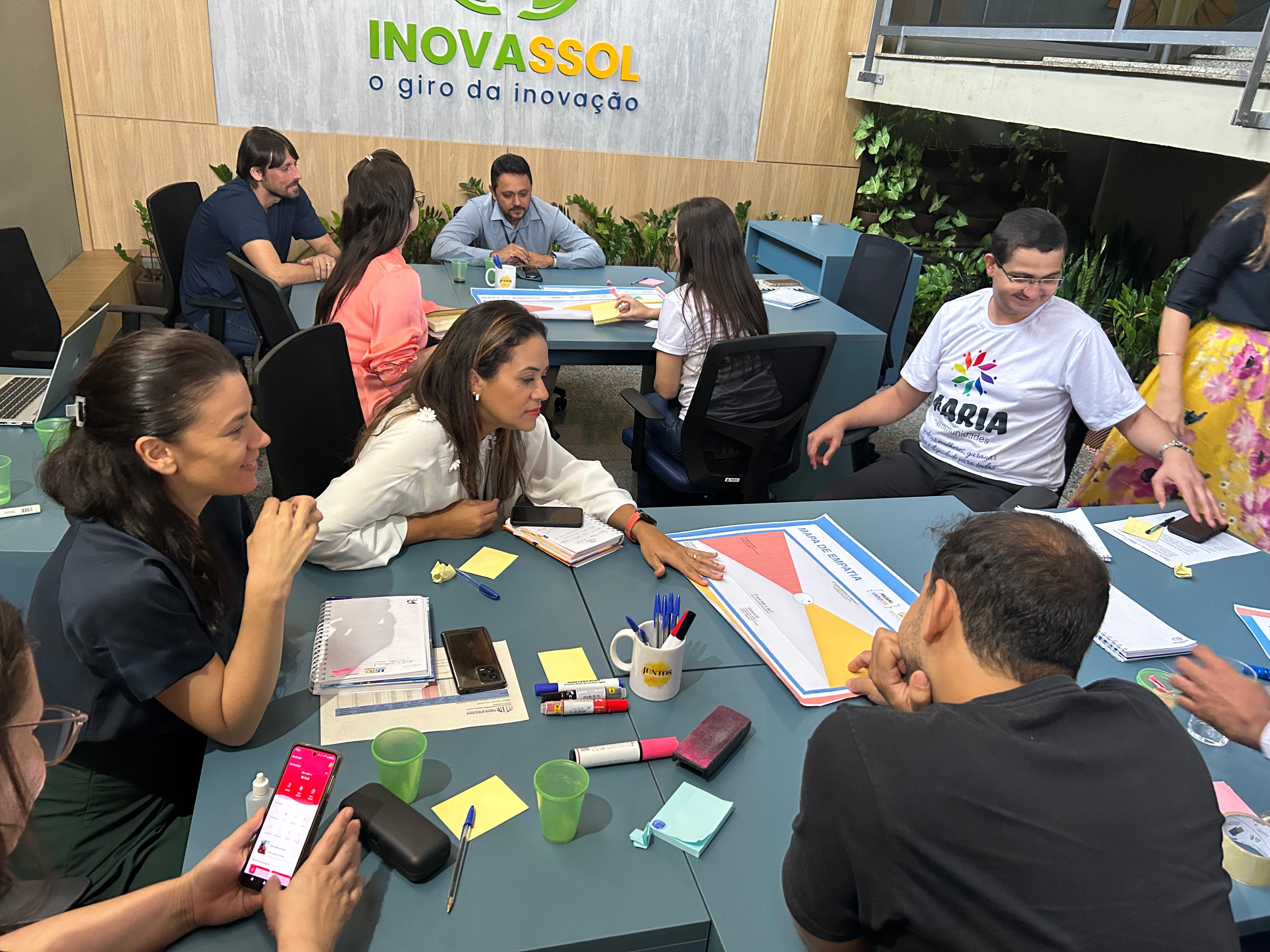 Fotografia colorida onde várias pessoas estão realizando atividades sentadas em volta de duas mesas separadas 