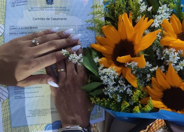 Fotografia colorida de duas mãos com alianças em cima de um documento de casamento, e ao lado um buquê de girassóis 