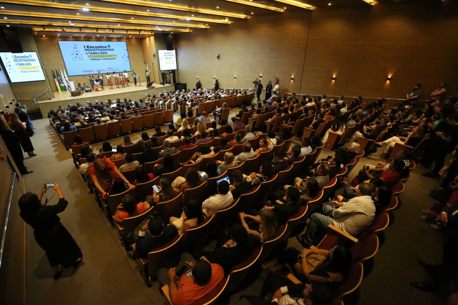 Auditório cheio de pessoas participando do evento e concentradas, com algumas pessoas de pé.