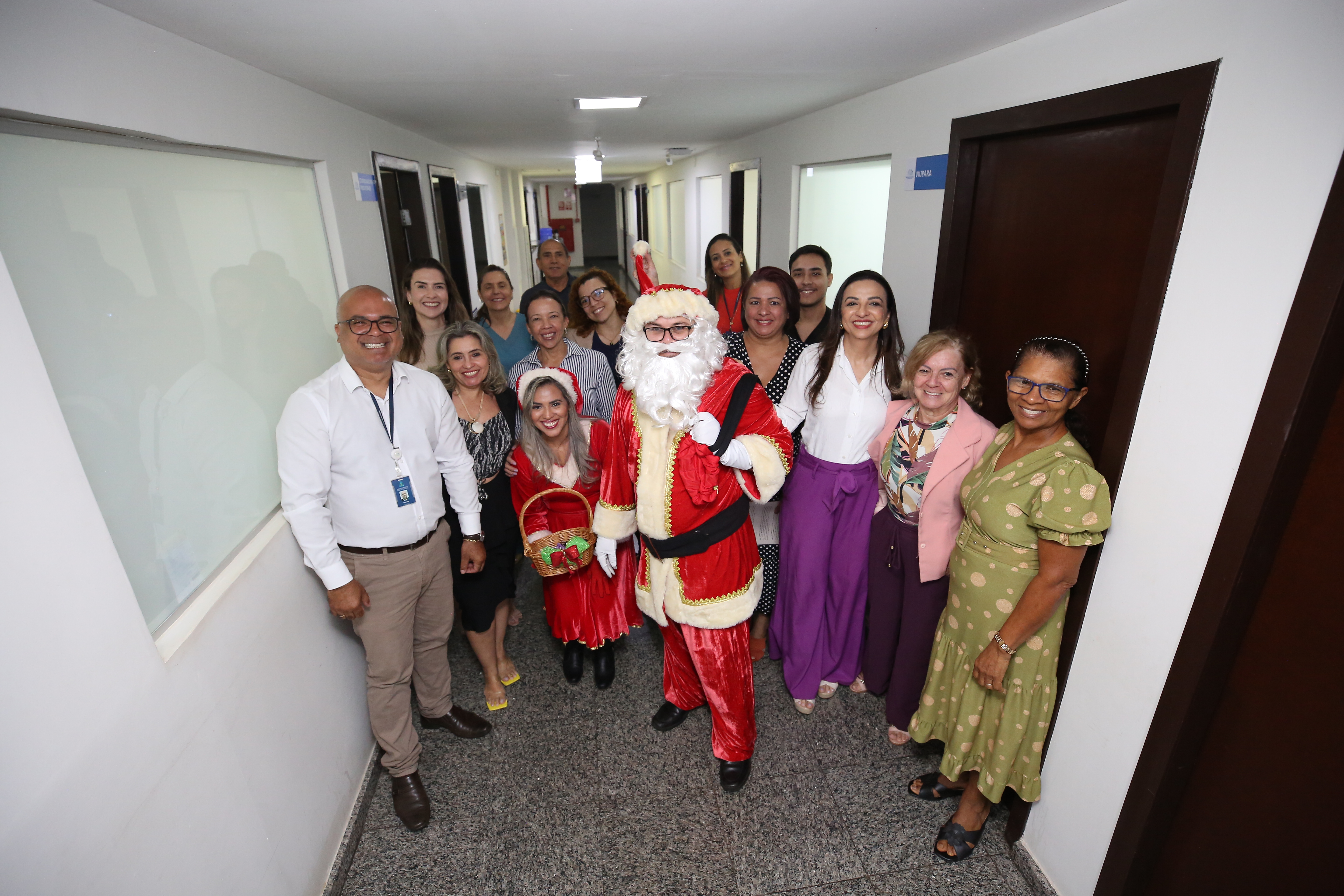 Fotografia colorida onde várias pessoas estão em um corredor repleto de janelas e portas de madeira e vidro. Ao centro desse grupo de pessoas, que posam para foto, estão um papai Noel e uma mamãe Noel 
