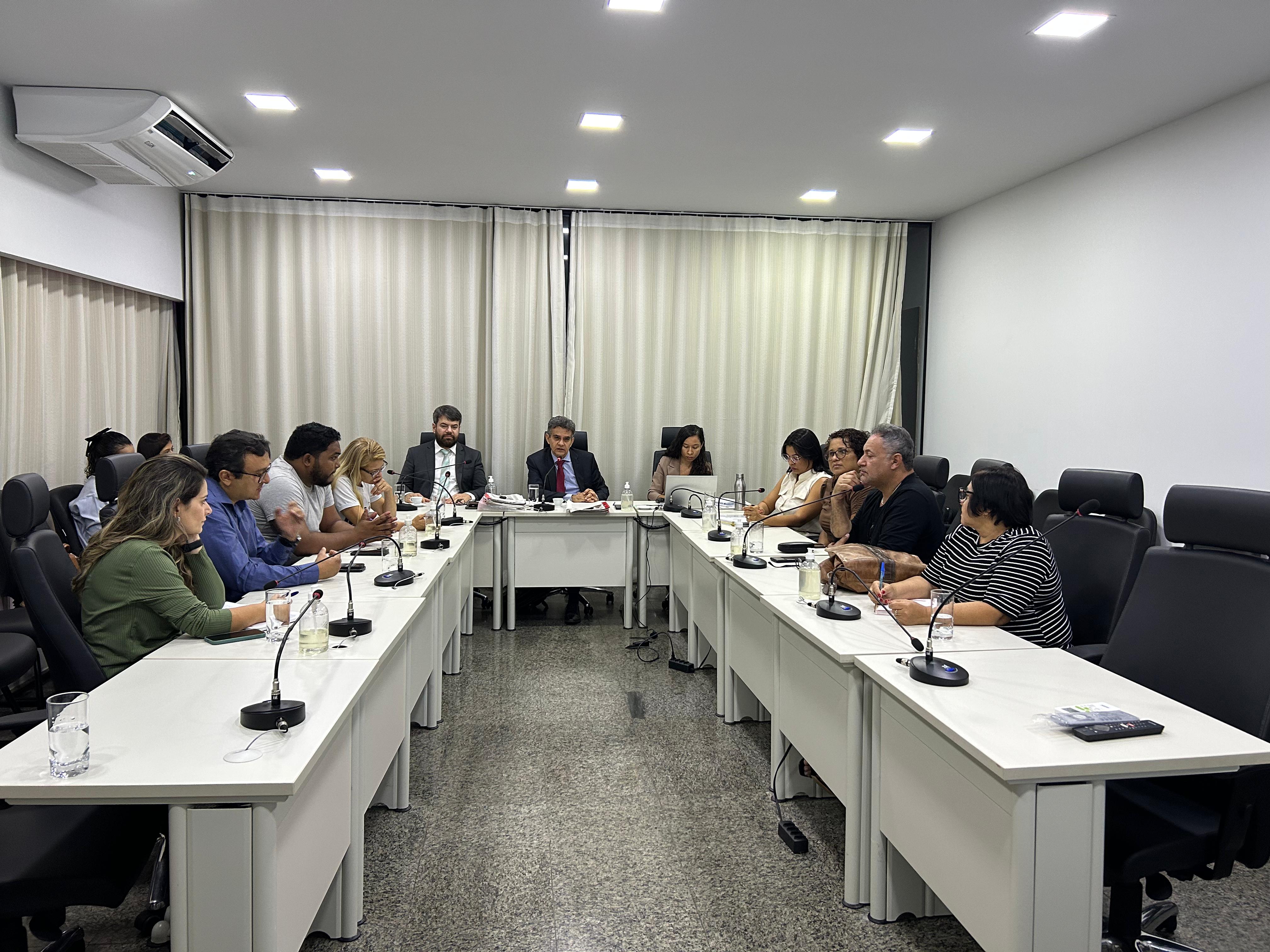 Fotografia colorida onde se encontra uma grande mesa de três partes, aberta no meio, com 11 pessoas sentadas em volta. Eles estão em uma sala branca com uma cortina bege ao fundo 