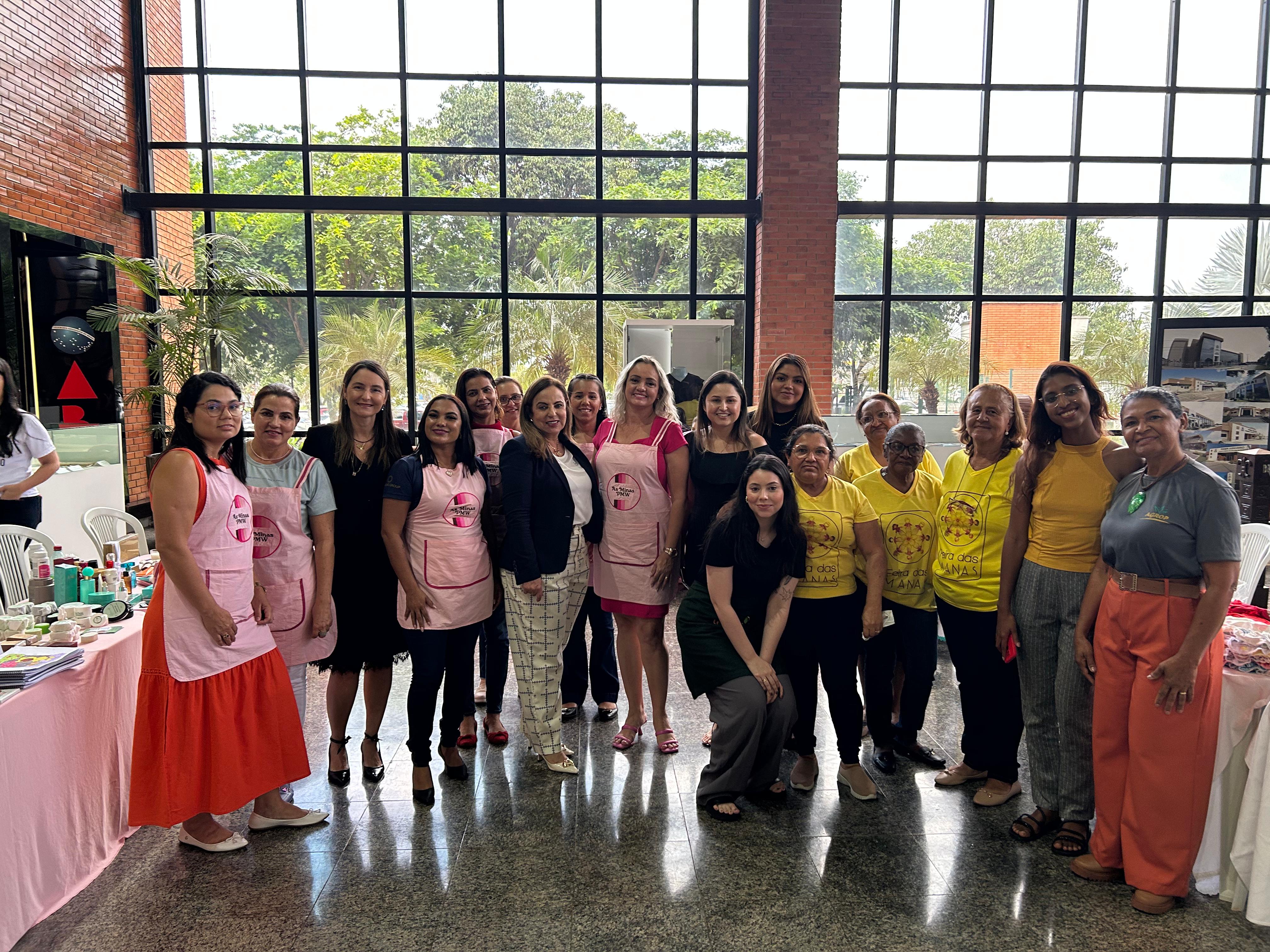 Fotografia colorida onde um grande grupo de mulheres está no hall principal do Tribunal de Justiça do Tocantins, posando para foto uma do lado da outra. Aos lados, tem mesas dispostas com forros coloridos e atrás uma grande janela de vidro que cobre toda a parede, dividida por várias linhas 