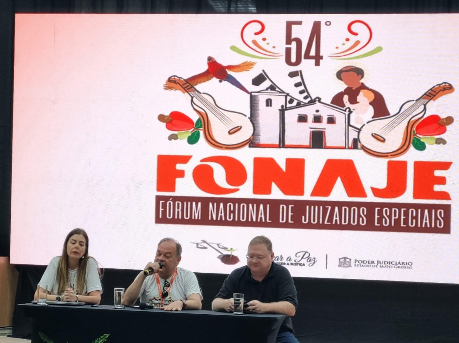 Fotografia colorida onde dois homens e uma mulher estão sentados lado a lado atrás de uma mesa preta com um telão branco de fundo, onde estão refletidos casas, instrumentos musicais e um Padre com uma criança. Abaixo, o nome do evento.