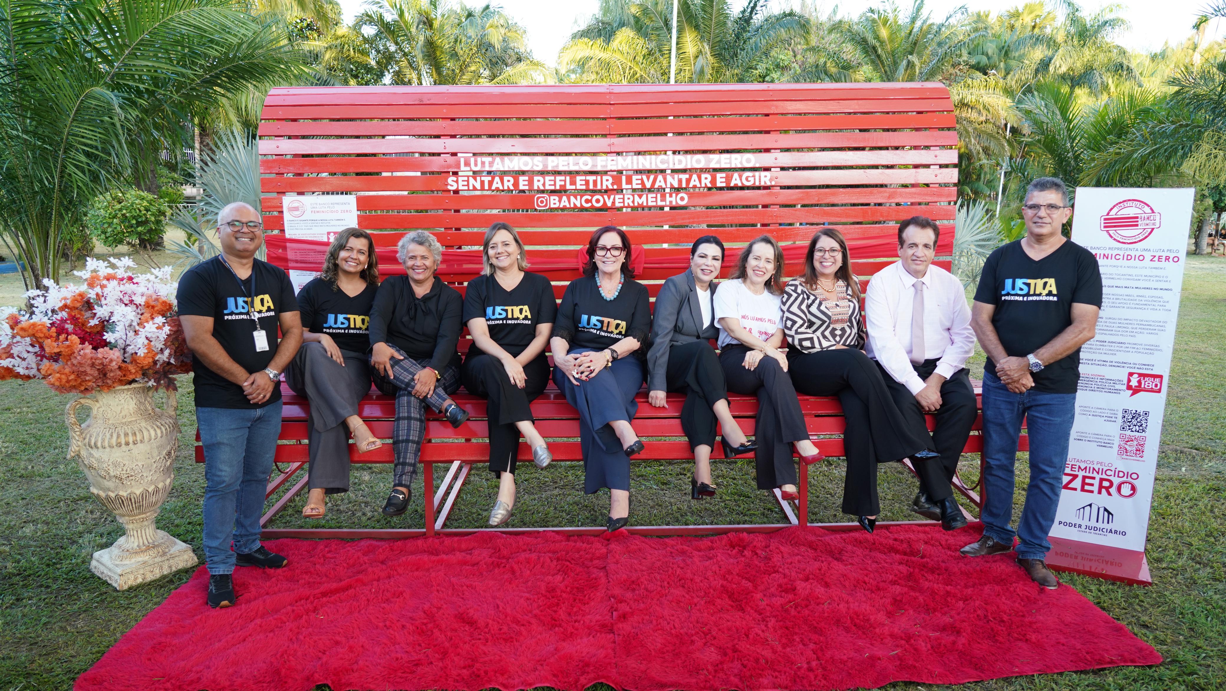 Fotografia colorida onde sete mulheres e um homem estão sentados em um grande banco vermelho, e em cada ponta do banco tem um homem de pé. Eles estão em um espaço aberto e no chão a grama verde toma conta, assim como um tapete vermelho em frente ao branco. De um lado do banco, junto de cada homem, tem um vaso com flores e um banner com algumas informações. 