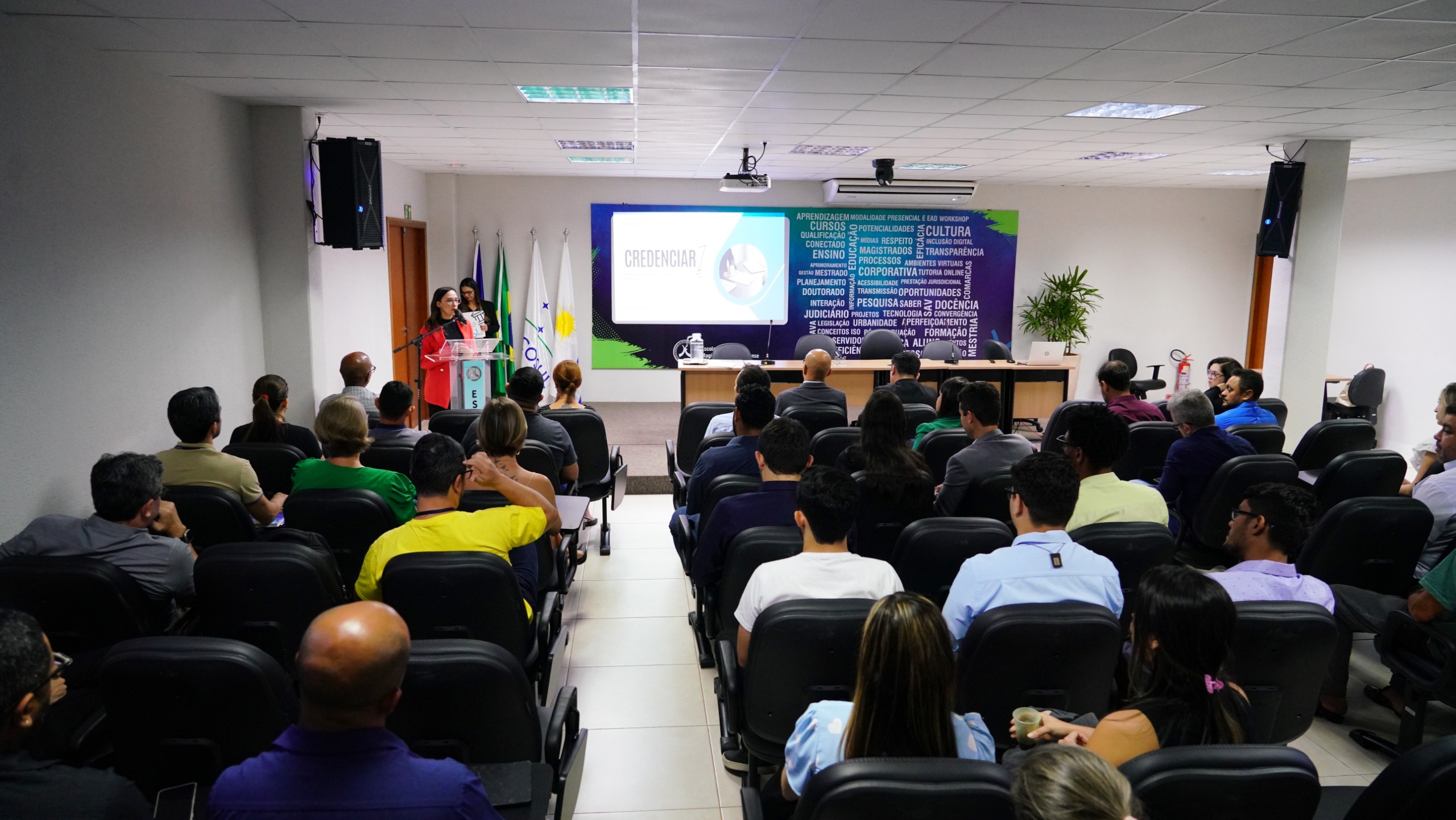 Fotografia colorida de uma reunião acontecendo em uma grande sala branca, com uma tela na parede a frente e atrás dela um mural azul cheio de palavras. Várias pessoas estão sentadas em cadeiras pretas, e a sua frente uma mulher de cabelos pretos e blazer vermelho palestra 