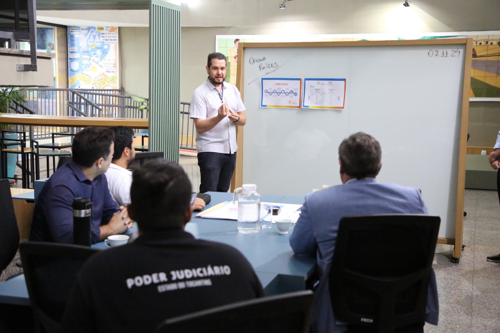 Fotografia colorida onde uma mesa com quatro homens que estão assistindo um outro homem ministrar uma atividade. Ele está de pé em frente a um quadro branco de vidro e conversa com os presentes na mesa. Eles estão em um local amplo e aberto, com medas, cadeiras e postas de vidro ao redor.