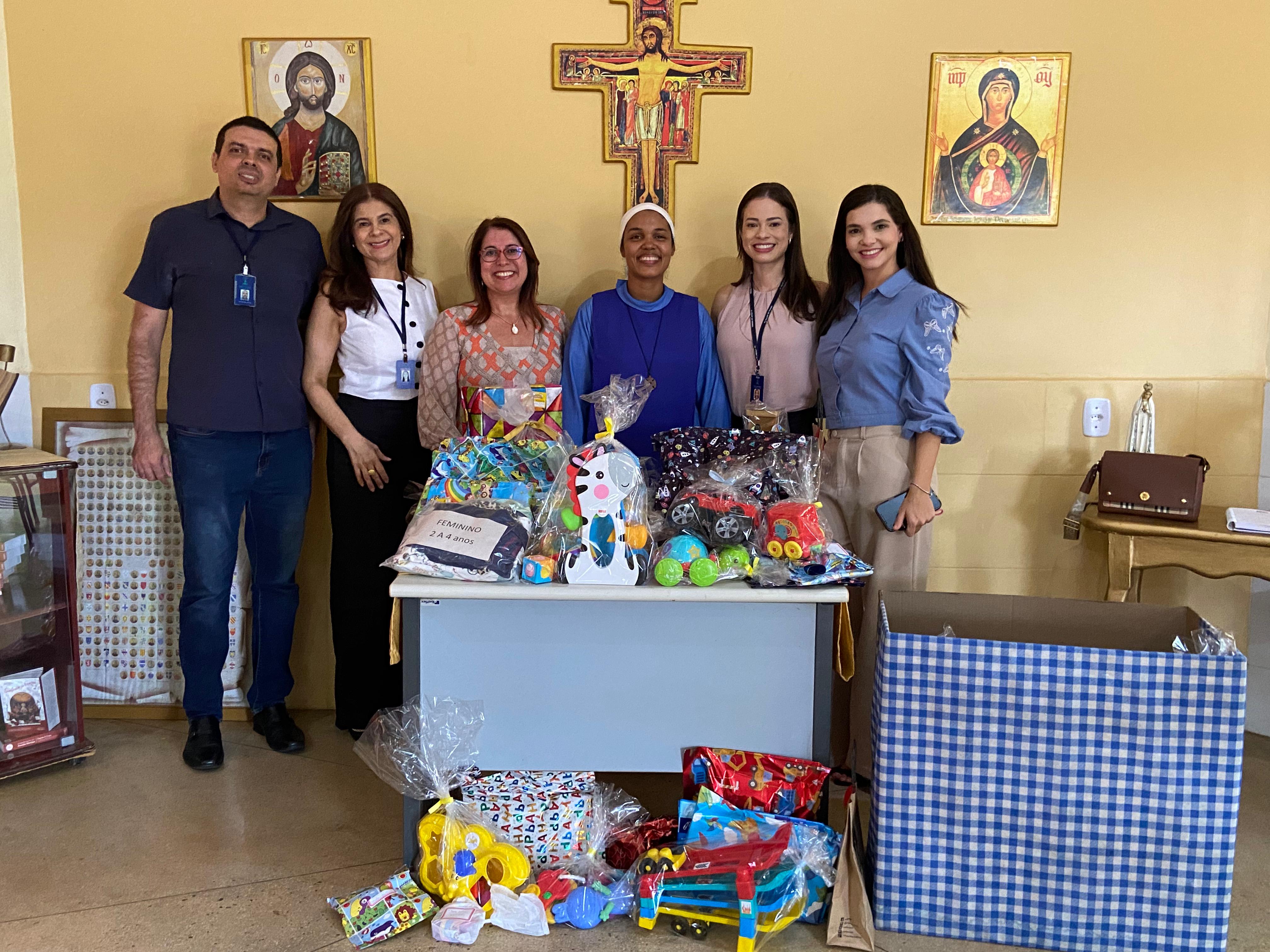 Fotografia colorida onde um grupo com cinco mulheres e um homem posam para fotografia lado a lado, em frente a uma mesa repleta de brinquedos embrulhados para presente, e uma caixa xadrez do lado. Um deles, a mulher do meio, é uma freira, e eles estão em uma sala com paredes amarelas com imagens religiosas nas paredes.