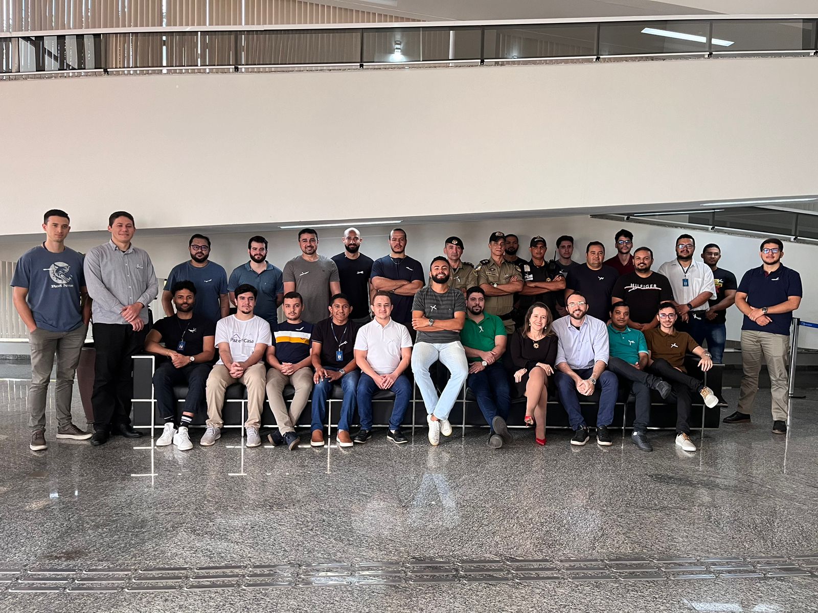 Fotografia colorida onde um grande grupo de homens está reunido em volta de um grande sofá preto. Alguns estão sentados e outros em pé. Em meio deles, tem uma mulher de vestido preto. Estão sentados em frente a lateral de uma rampa de cor bege.