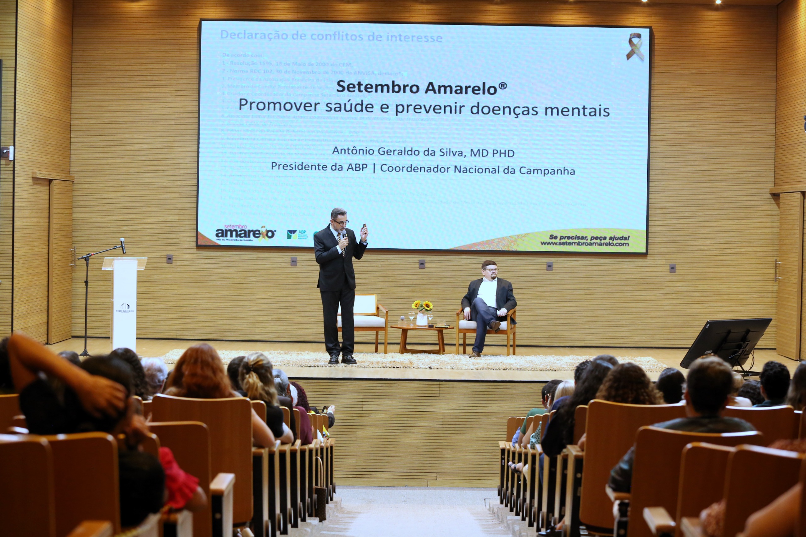 Homem de roupa social em cima de um palco de madeira clara com um telão atrás e outro homem sentado ao seu lado enquanto uma plateia assiste a frente 