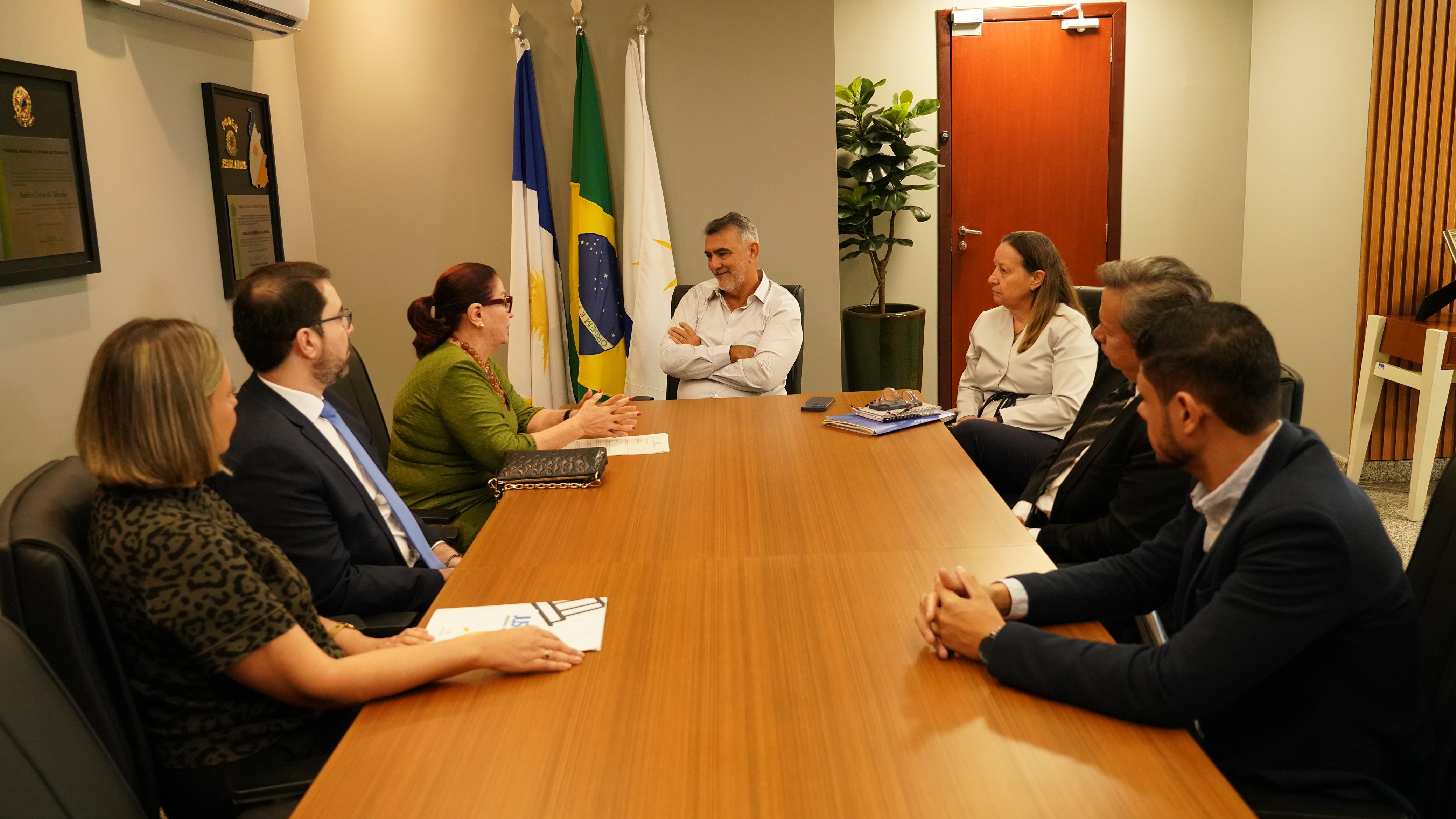 Desembargadora Etelvina Maria Sampaio e demais integrantes do Poder Judiciário do Tocantins reunidos com o presidente da Assembleia Legislativa; eles estão sentado em uma mesa grande de reuniões, ao fundo uma porta de madeira e um conjunto de bandeiras do Tocantins, Brasil e  outra que não dá ara identificar