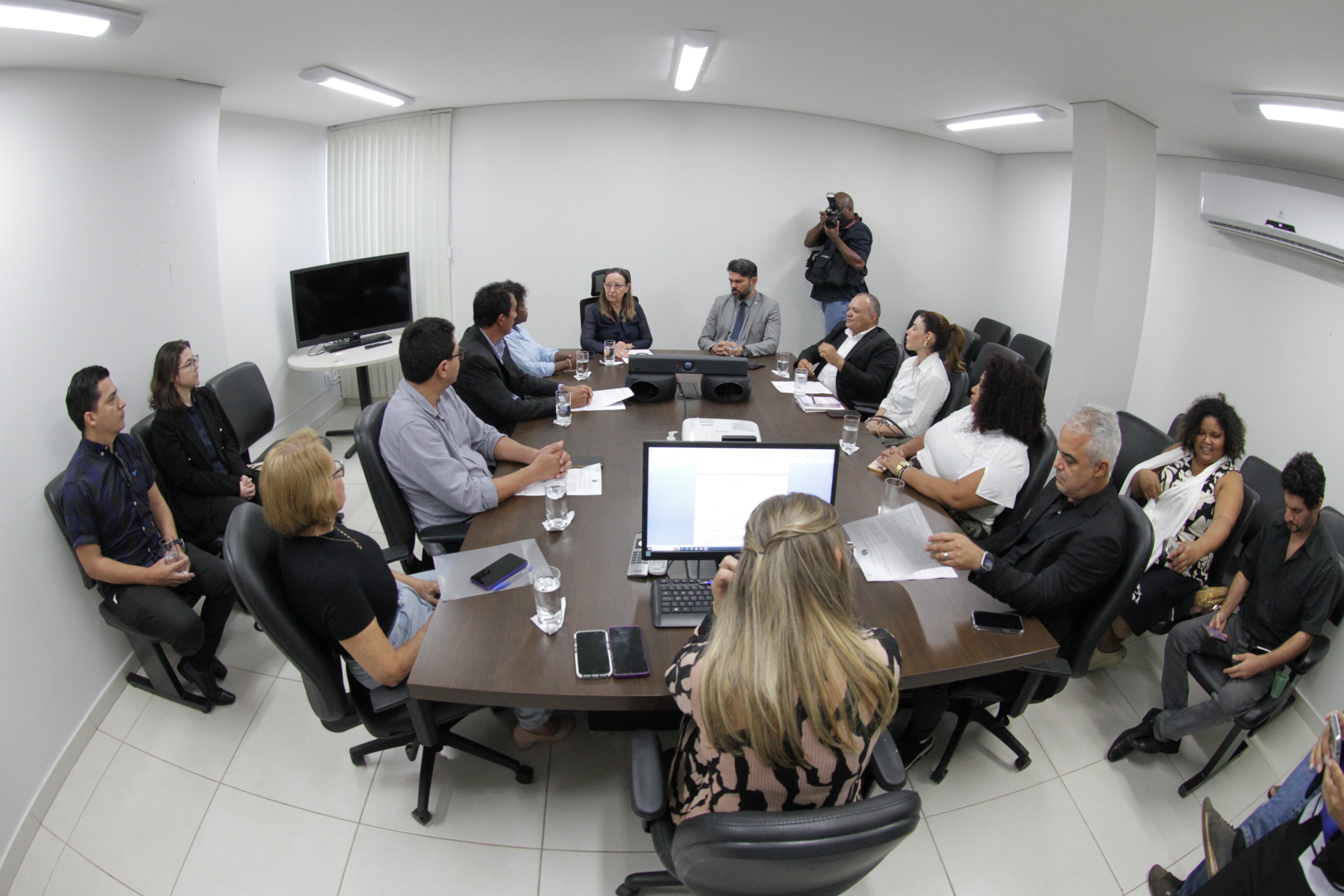 Grupo de pessoas sentadas ao redor de uma mesa retangular, algumas com papéis à sua frente.  A sala é iluminada com luz branca e o ambiente é corporativo, com paredes claras e mobília de escritório.