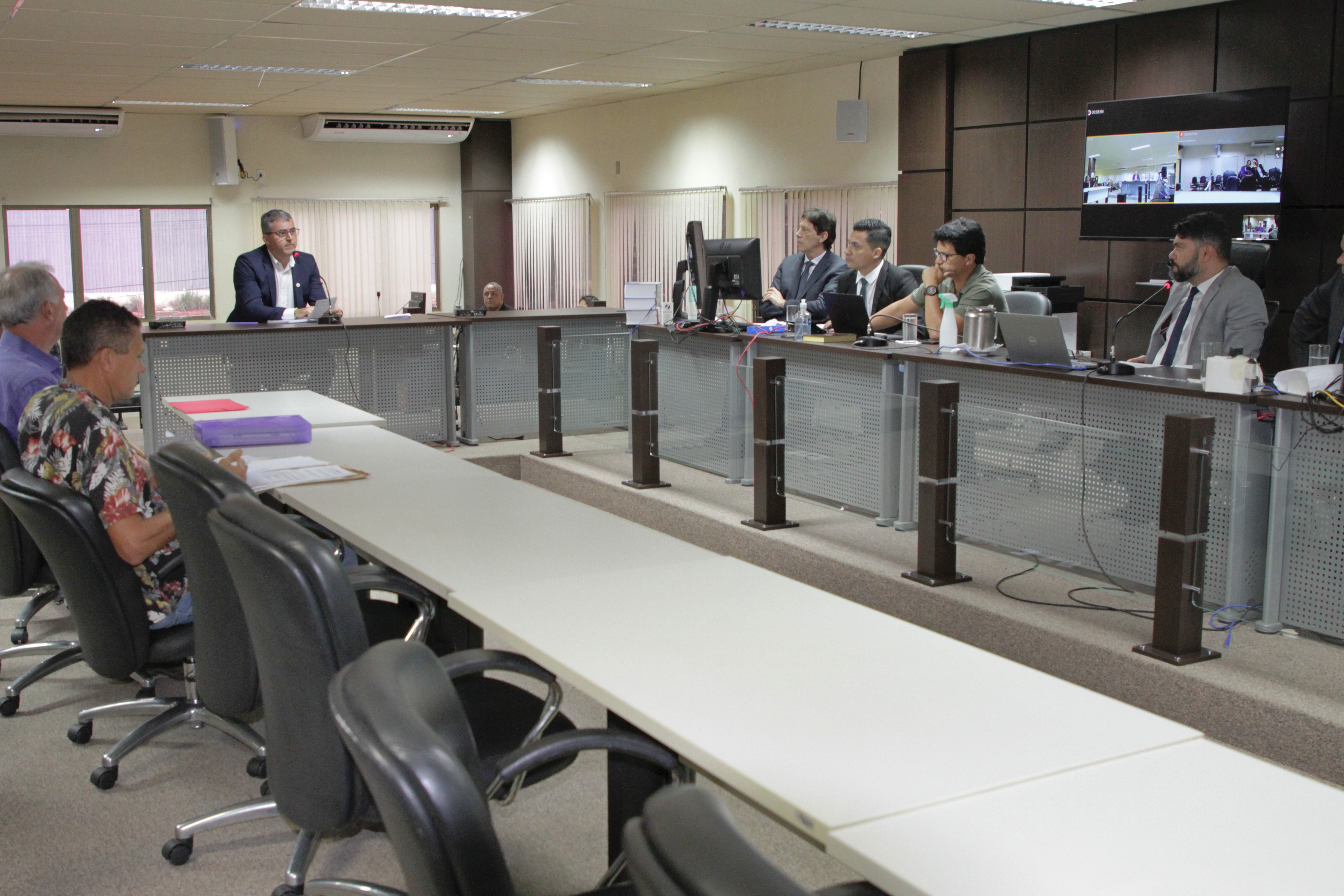 Grupo de pessoas sentadas ao redor de mesas e uma grande tela na parede exibe uma videoconferência. O ambiente é bem iluminado.