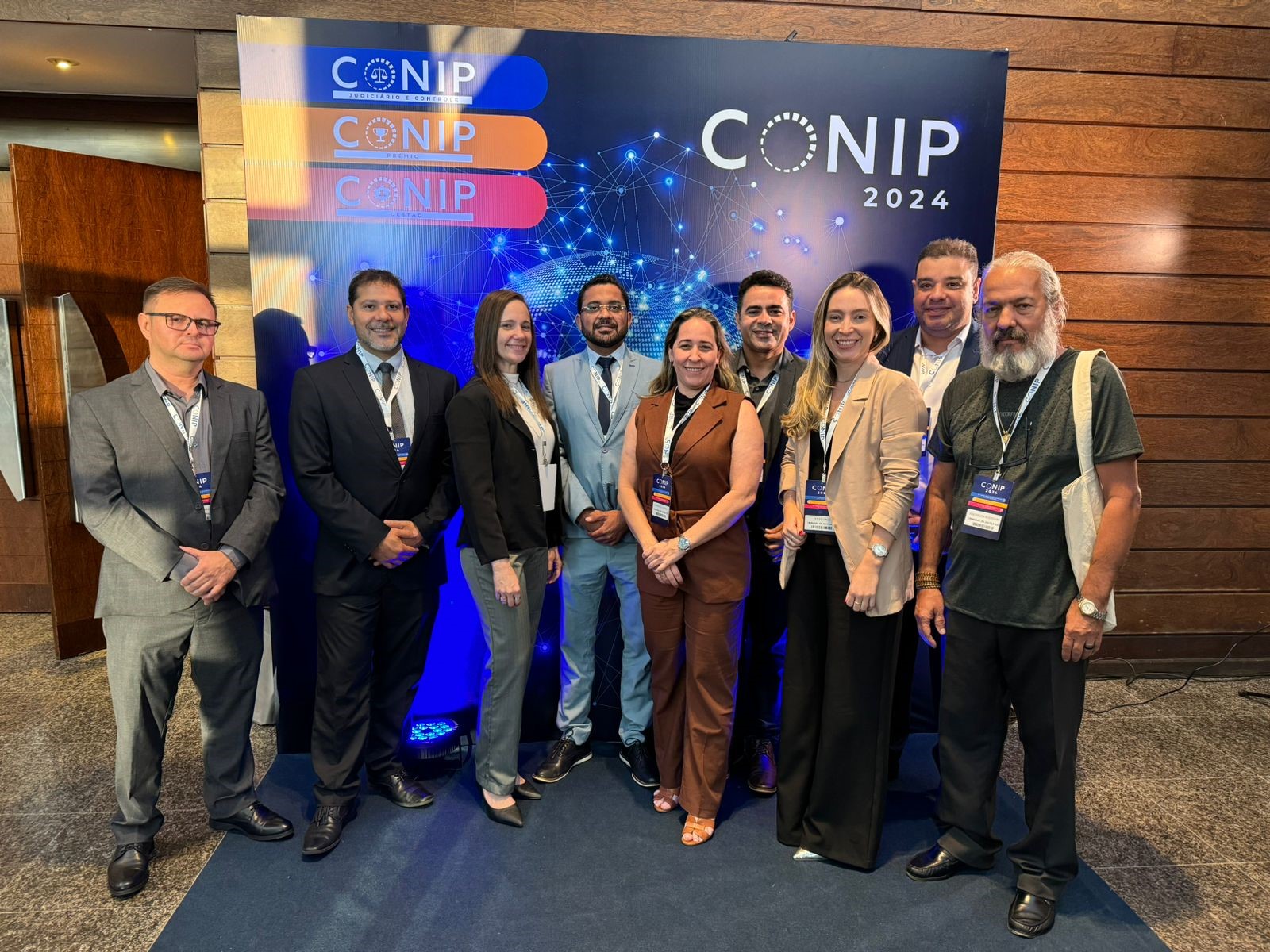 Grupo de nove pessoas vestidas formalmente, todos em pé frente ao banner com os dizeres "Conip 2024'
