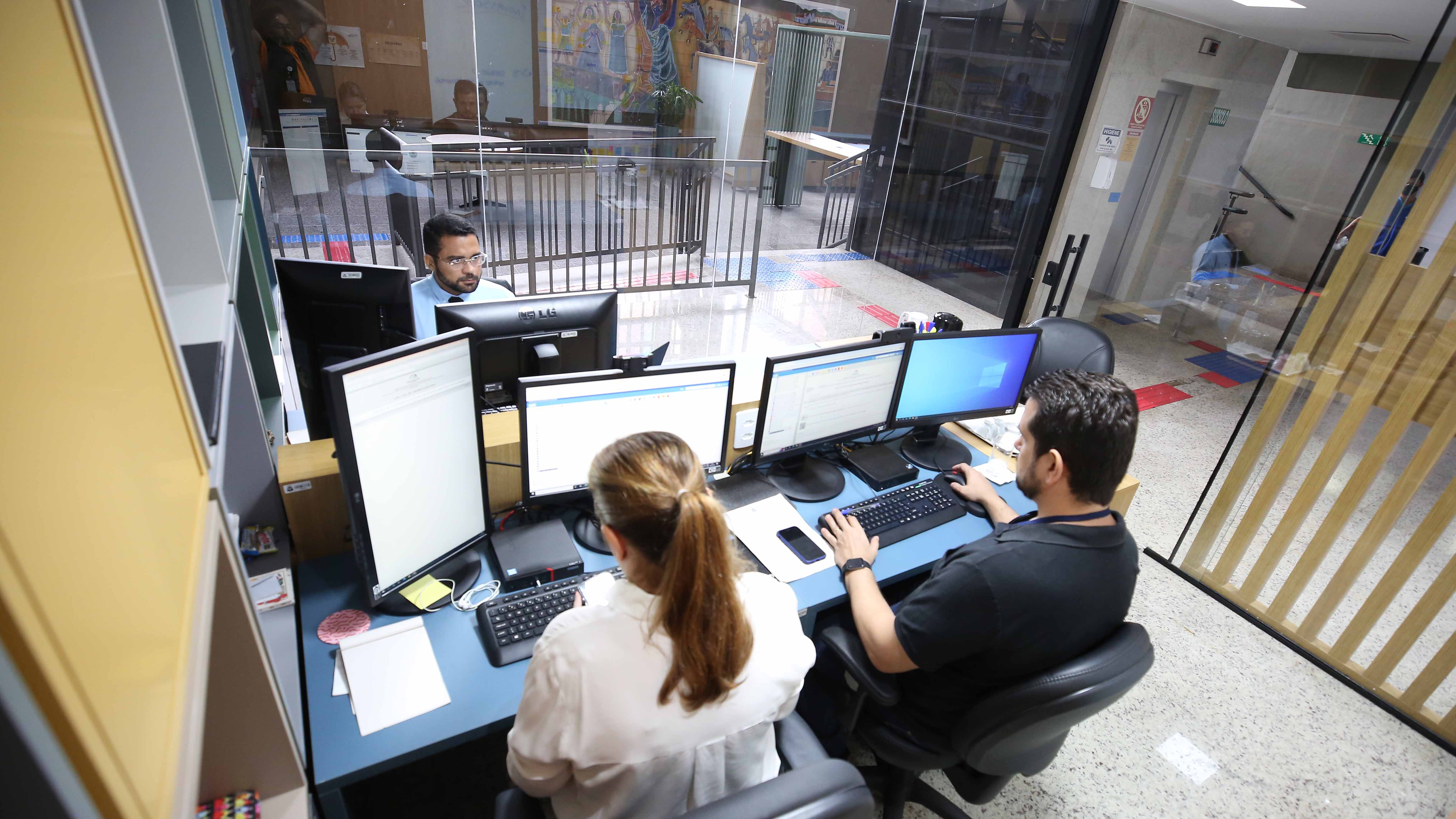 Imagem colorida que mostra três pessoas, sendo dois homens e uma mulher, trabalhando em frente a computadores em uma sala decorada