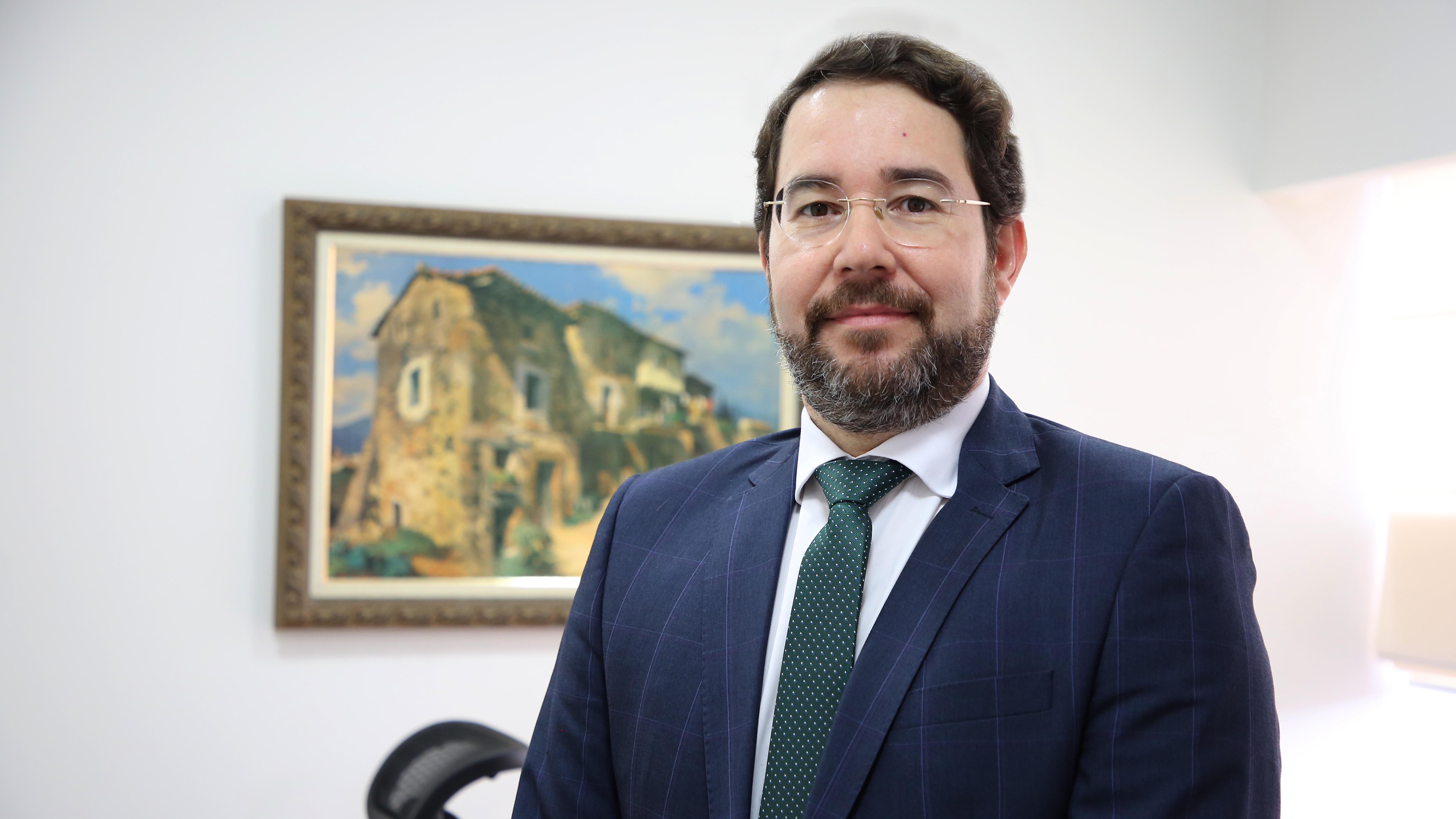 Dr. Roniclay, homem de cabelos escuros, barba e óculos; ele usa terno azul escuro, camisa branca e gravata verde escuro