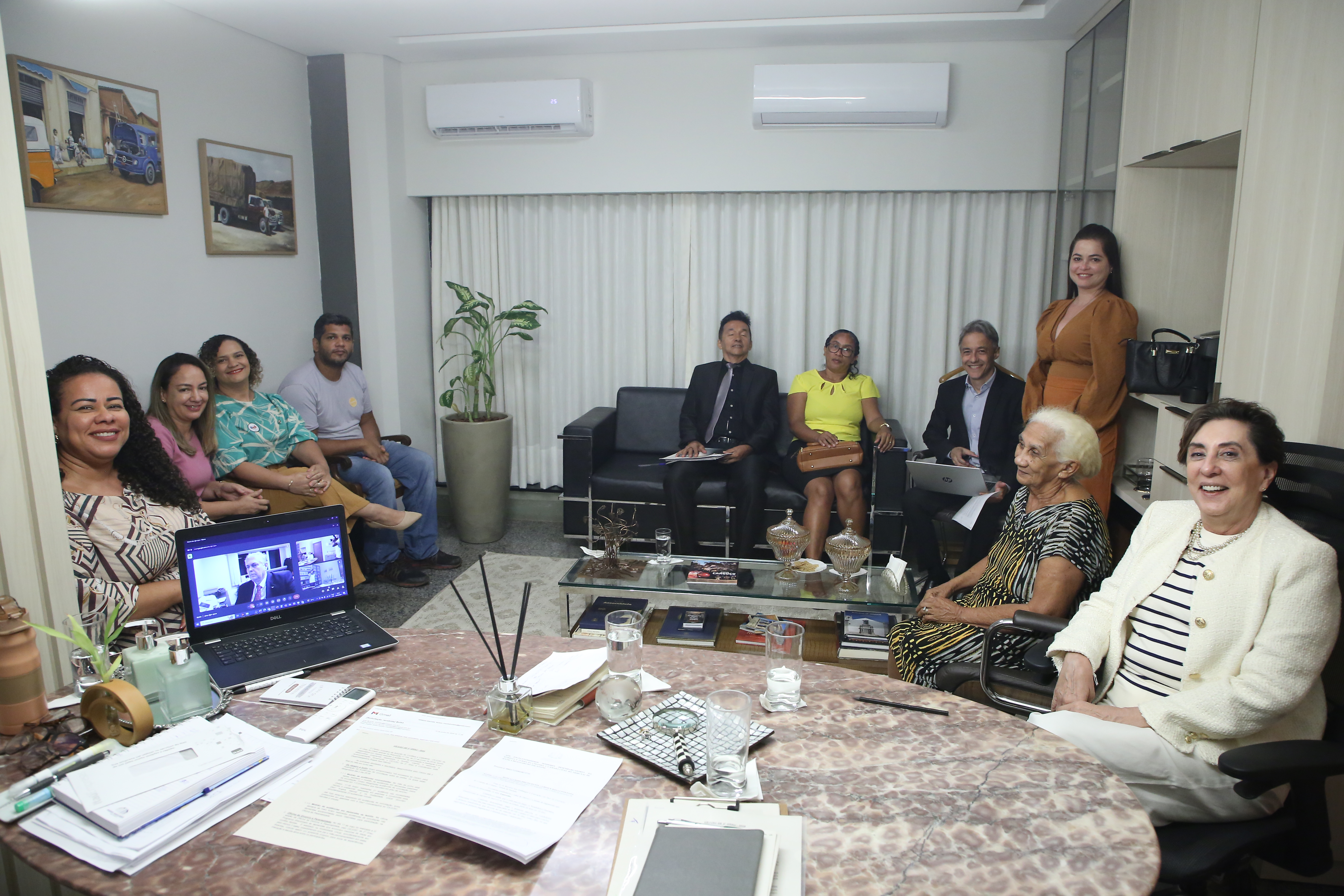 Imagem aberta do gabinete da desembargadora Angela Haonat; na foto, direita para a esquerda estão: a desembargadora (de terninho branco e blusa com listras pretas e brancas), uma mulher idosa de cabelos brancos, uma conciliadora (em pé, de vestido marrom), um assessor (de cabelos grisalhos, terno escuro e camisa azul, com um notebook nas pernas; uma mulher de óculos, blusa amarela, saia preta e uma bolsa nas benas; um homem de roupa preta e gravata cinza e pasta com documentos nas pernas; um home moreno de camiseta cinza e calça jeans; e três mulheres sentadas na sequencia.