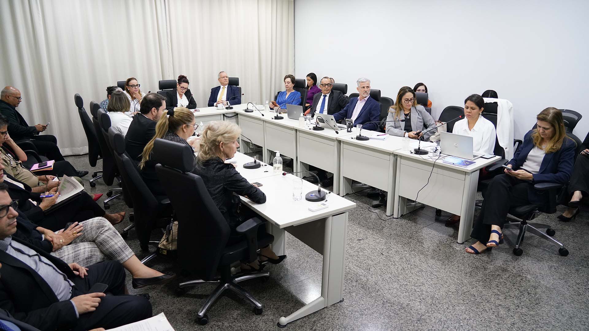 Desembargadores, juízes, diretores, coordenadores e assessores reunidos em uma sala de reuniões; na imagem aparece mesas brancas e formato em "u" e os participantes sentados, alguns com notebooks, discutindo pautas da reunião
