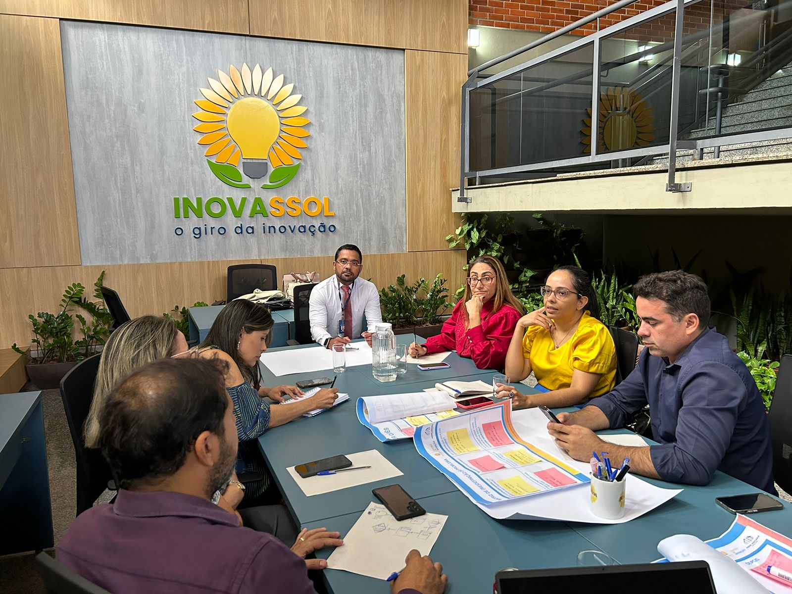 Quatro mulheres e três homens sentados ao redor de uma mesa azul, ao fundo a logo do Centro de Inovação - Inovassol