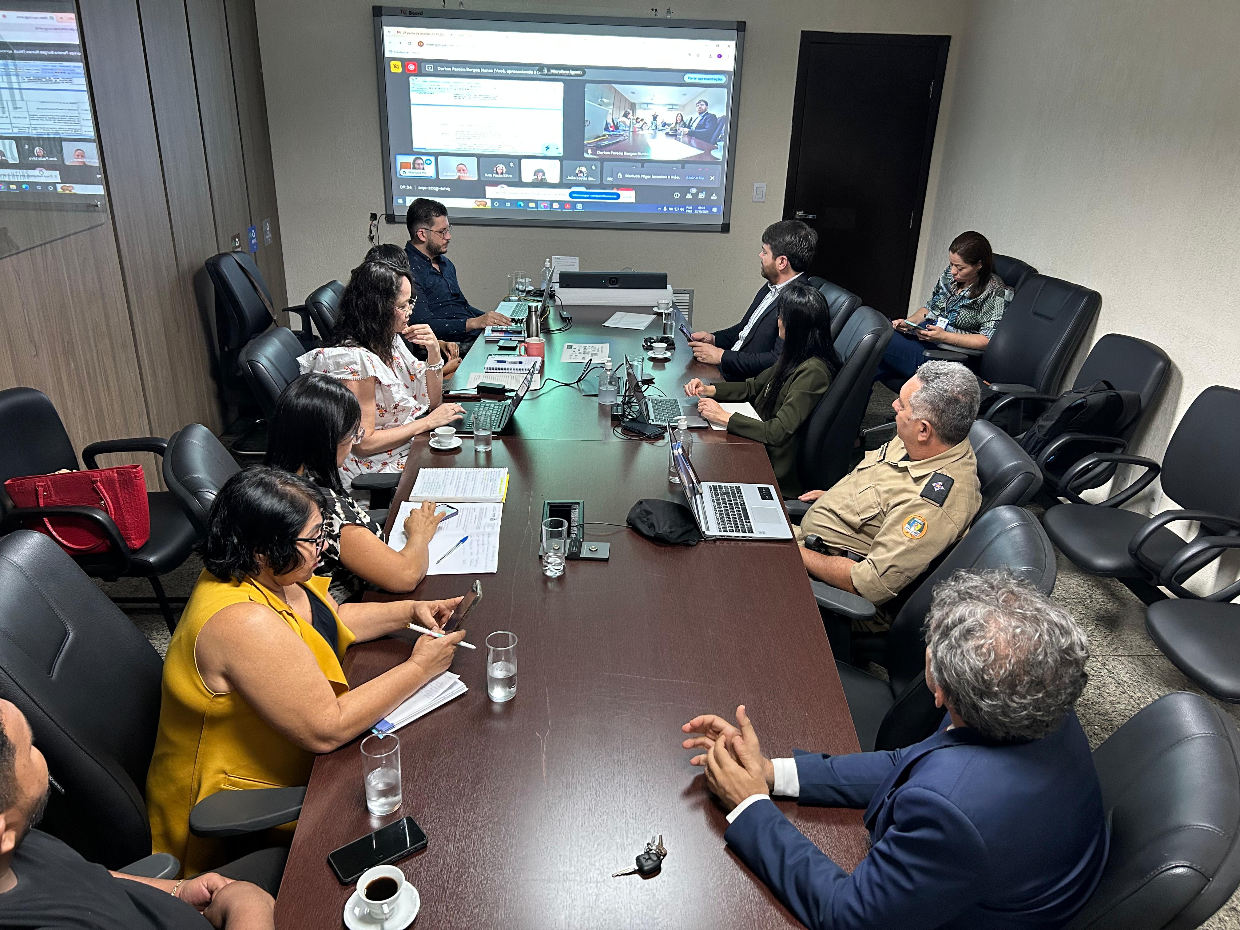 Sala de reuniões com várias pessoas reunidas em volta de uma mesa; ao fundo aparece um monitor grande onde está sendo projetada uma imagem e todos voltados para lá