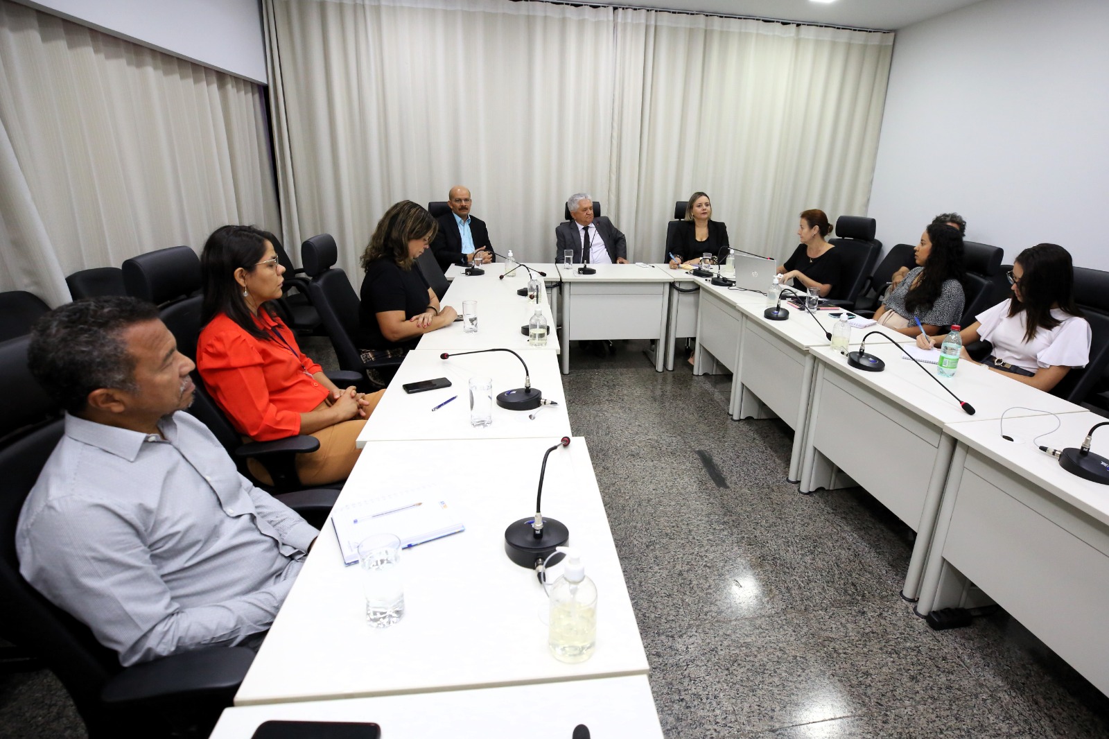 sala de reunião com desembargador, diretores e servidores do TJ reunidos para tratar sobre o concurso de juiz substituto; sala tem mesas brancas e cortinas e paredes brancas, cadeiras pretas